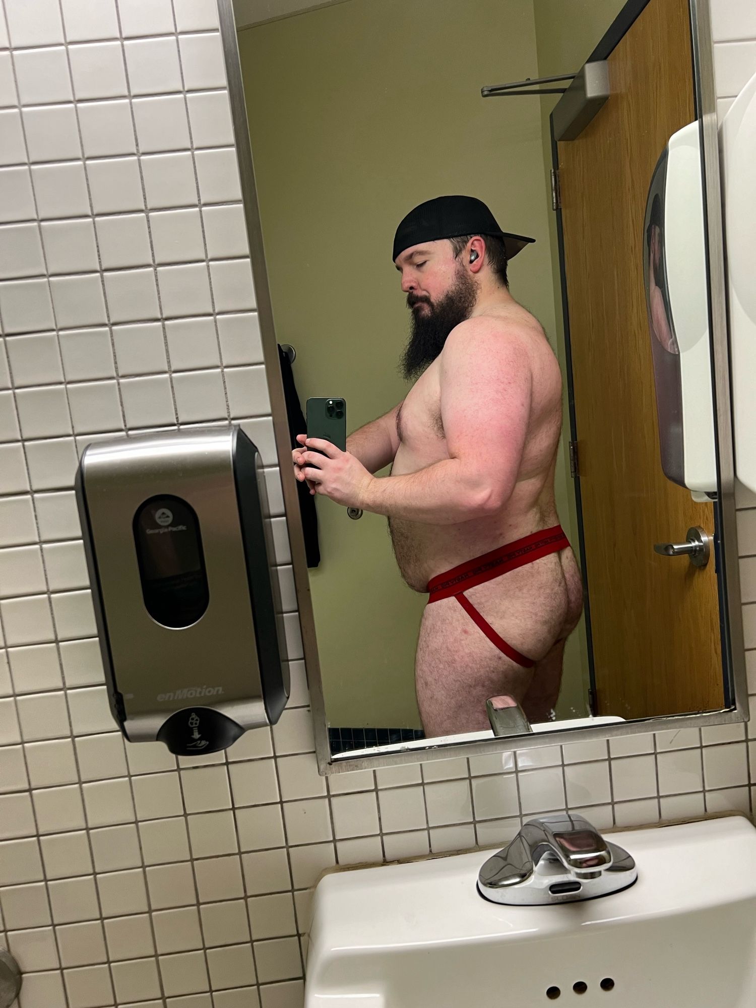 a bearish man wearing a backward black baseball cap and a red jockstrap taking a picture of his back and butt in a changing room mirror