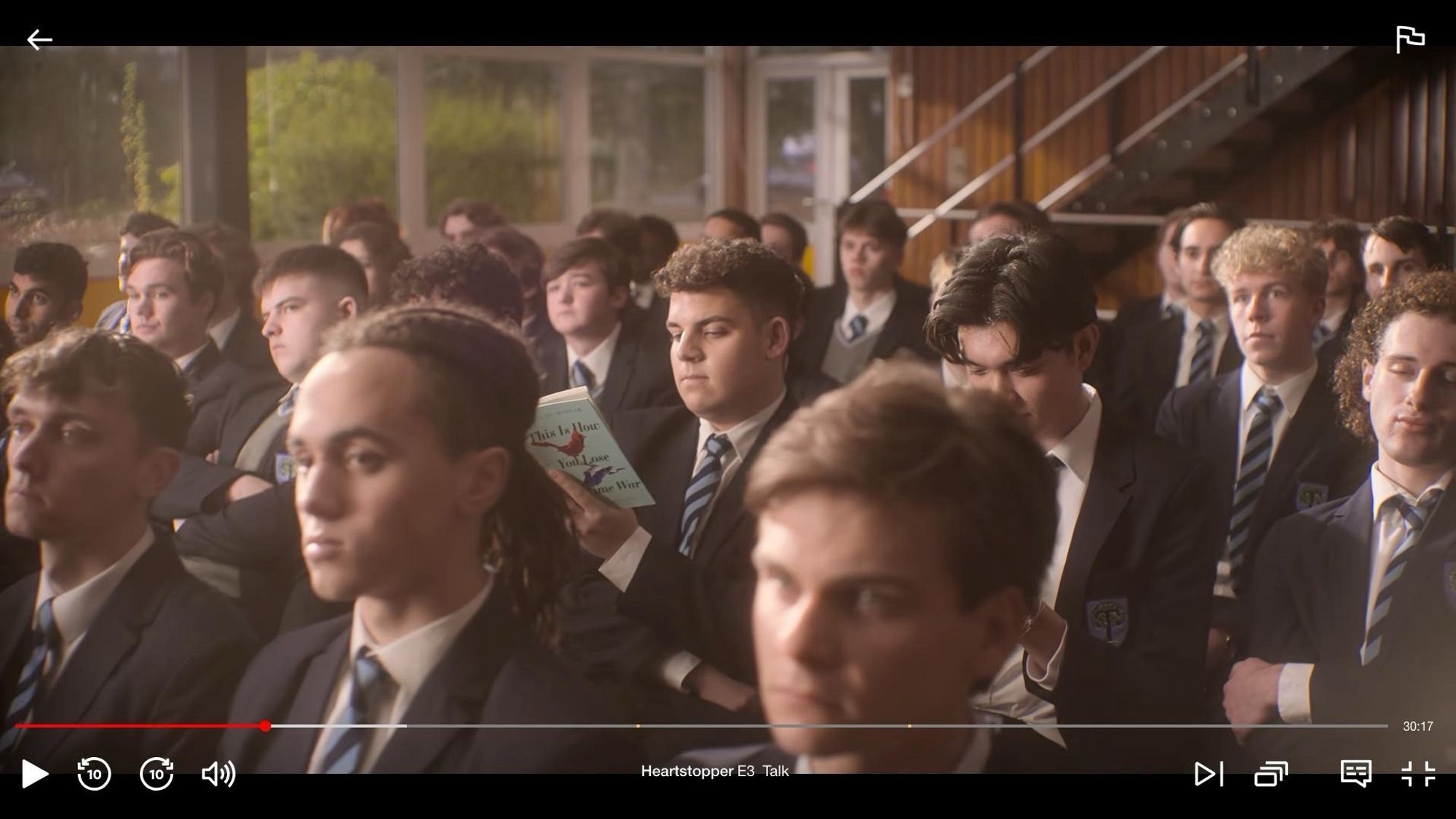 Screenshot of a crowd scene from S3 of HEARTSTOPPER in which a boy is reading a copy of THIS IS HOW YOU LOSE THE TIME WAR, a book I wrote half of!