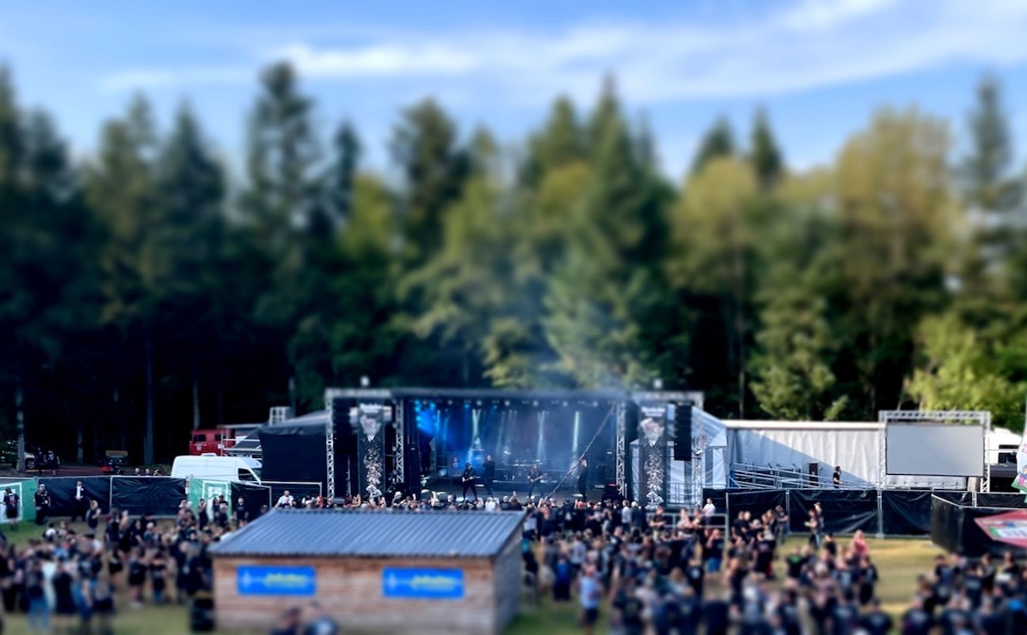 Das Metalacker-Festival (2023) in einer Tiltshift-Aufnahme: Die Bühne am Waldrand und Publikum. Es dürfte so früher Nachmittag sein.