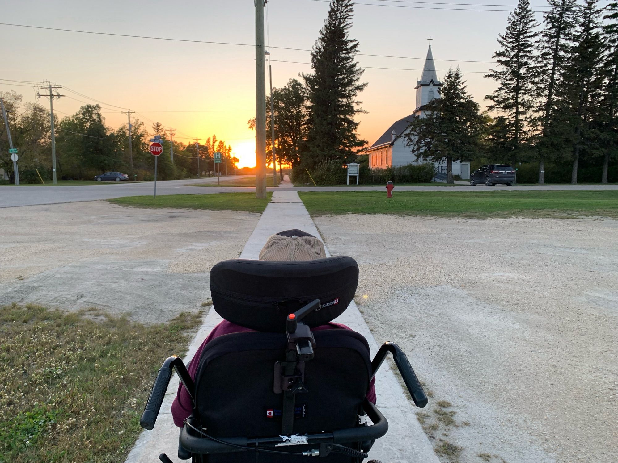 Pushing Dad in his wheelchair down a paved sidewalk at dusk, leading to a glowing sunset. There’s an old wooden church on the right, surrounded by tall spruce trees.