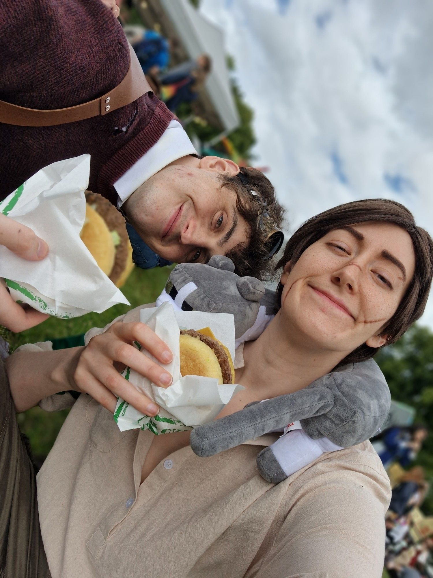 cosplayers of Grian and Scar, each with a burger in hand, smiling, 'Grians' head is leaning on 'Scars' shoulder