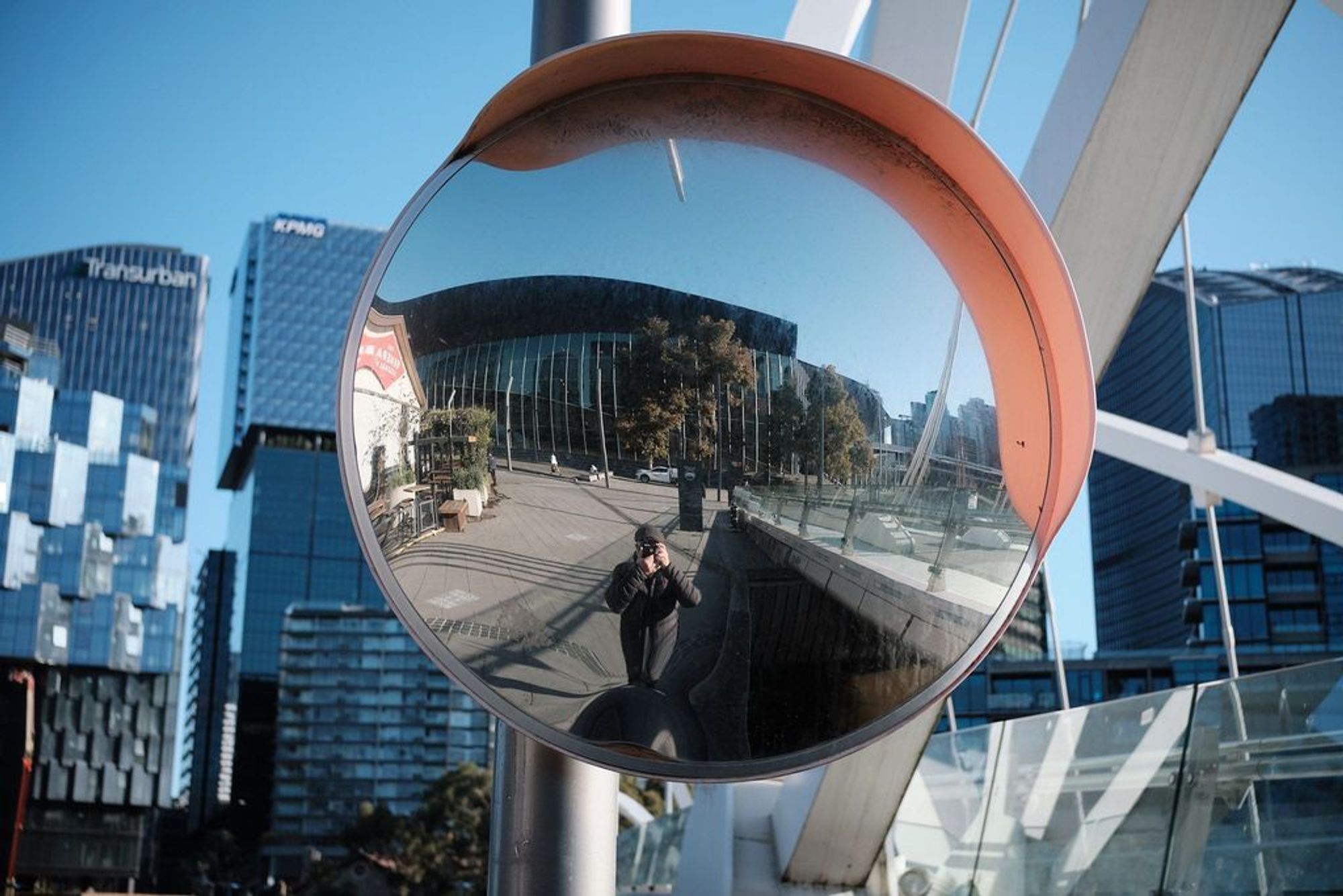 Close-up photo of a large, convex, traffic safety mirror installed on a pole in a blind curve along a cycling and walking path next to an urban river. The photographer has captured his own distorted reflection in this mirror.