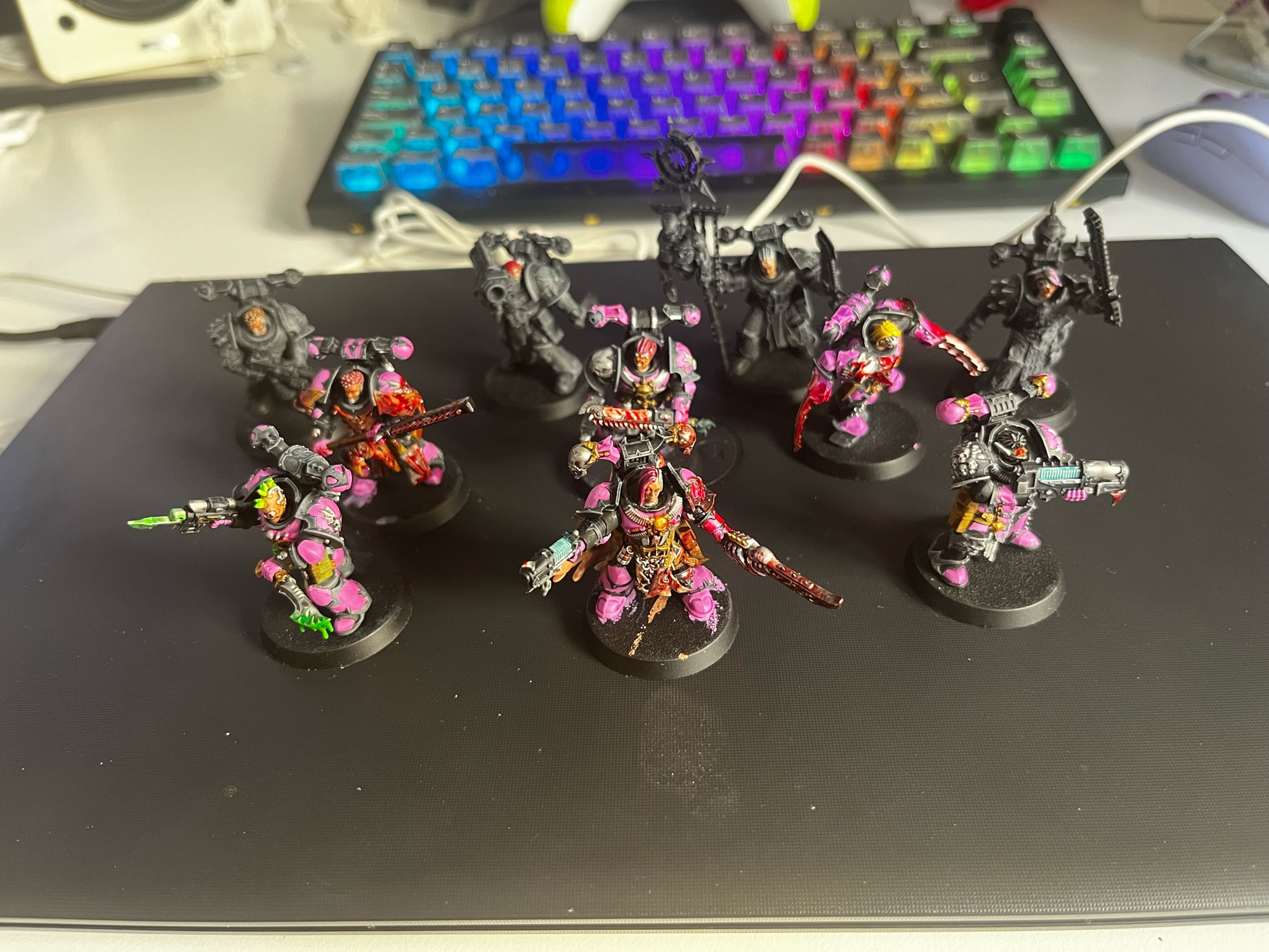 10 miniaturas de plástico del juego Kill Team en proceso de pintura, 6 de ellas casi terminadas y 4 apenas empezadas. Llevan armadura de color rosa, una variedad de armas de ciencia-ficción y salpicaduras imitando sangre por todas partes.