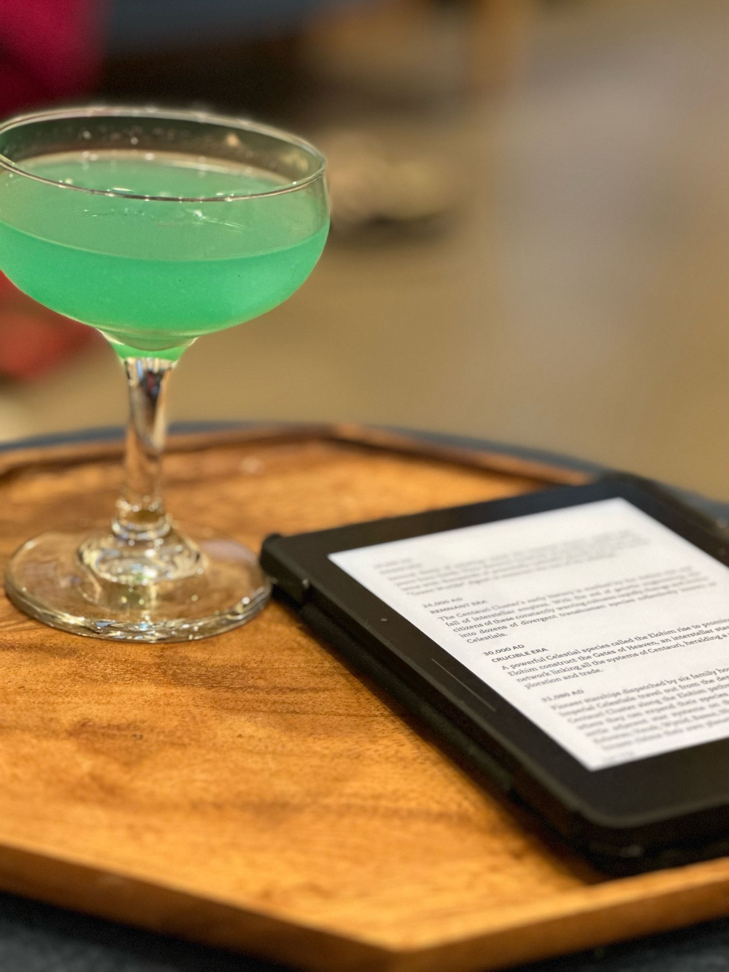 The image shows a light green cocktail in a coupe glass placed on a wooden tray. Next to the glass is an e-reader displaying a text document or book, placed flat on the tray. The background is softly blurred, with indistinct objects and warm lighting, creating a relaxed and cozy ambiance. The focus is on the drink and the e-reader, suggesting a quiet, leisurely moment of reading with a refreshing beverage.