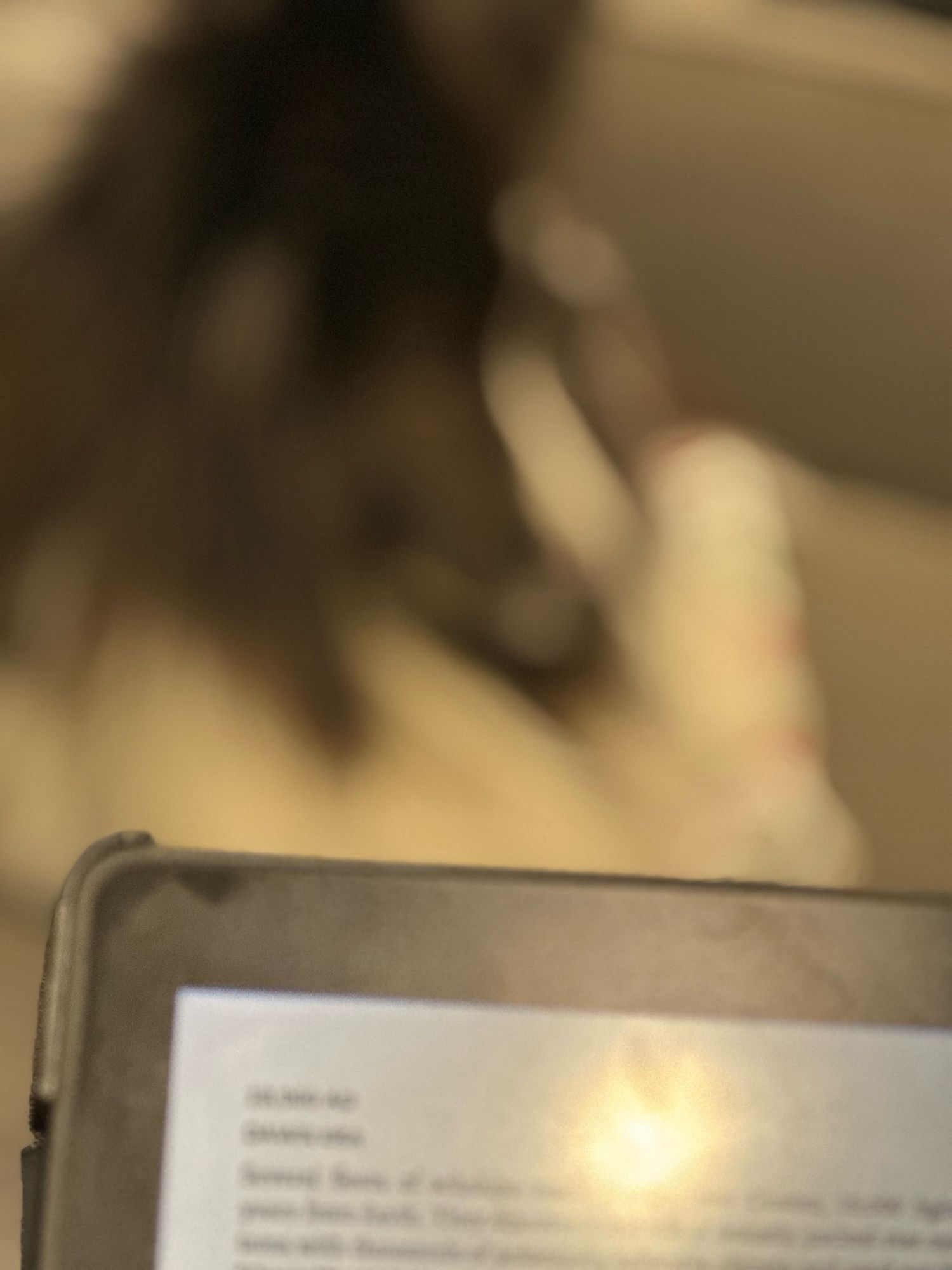 The image shows a close-up of an e-reader in the foreground, slightly out of focus, displaying text on its screen. In the blurred background, a dark-colored dog is lying on a couch, only partially visible due to the shallow depth of field. The scene suggests a relaxed setting, possibly someone reading while their pet rests nearby. The soft lighting adds to the cozy atmosphere.