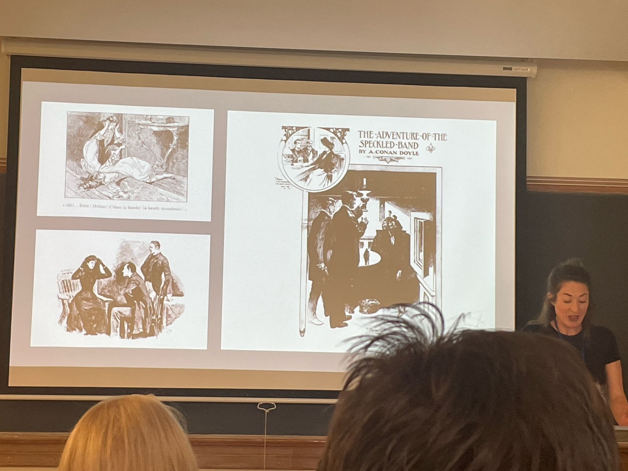 A woman at a podium {lectern} at the right of of the frame. Images of illustrations on slide projection.