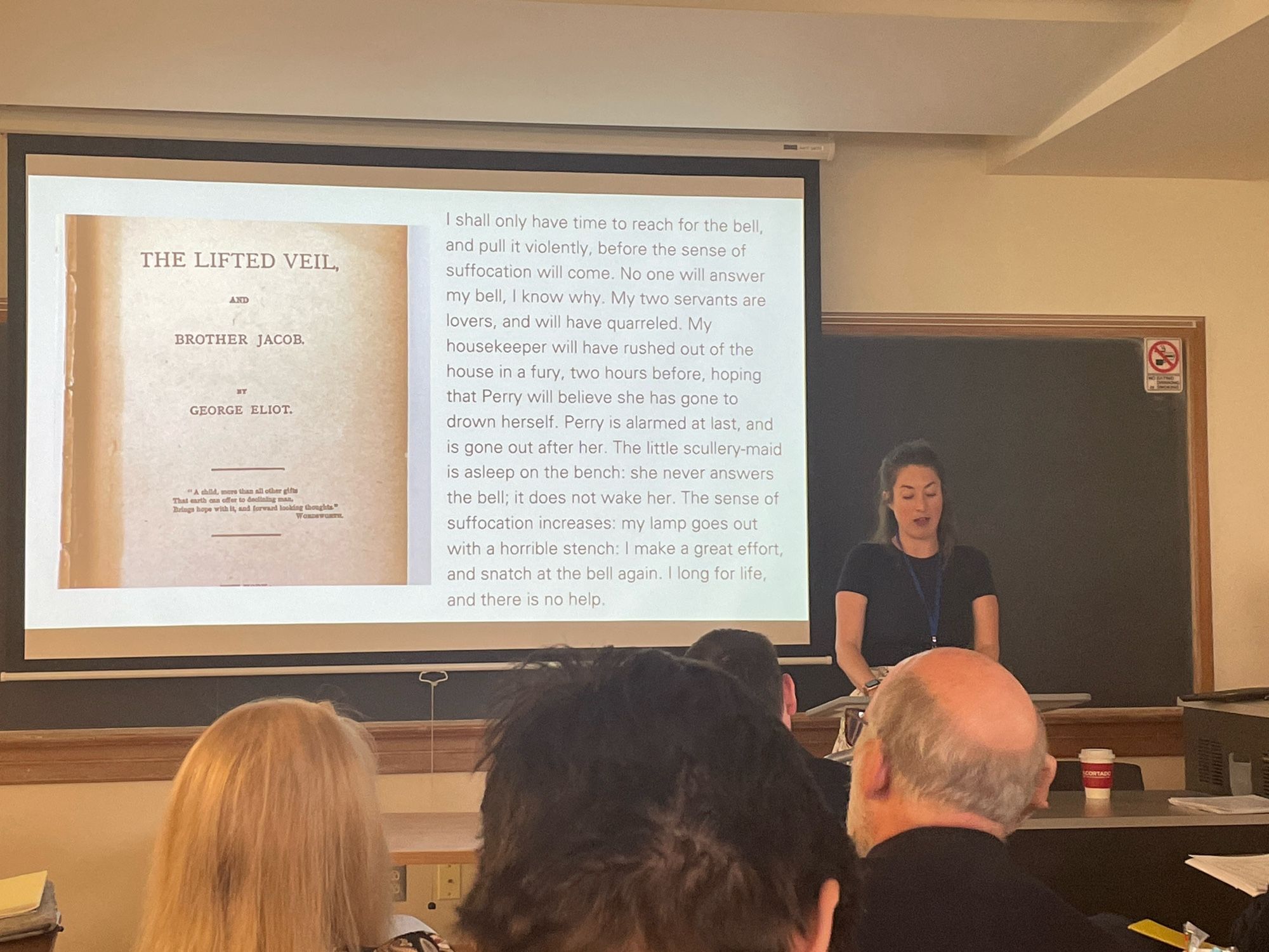 A woman stands at a podium {lectern} on the right of the frame. Slide projection shows a title page and text.