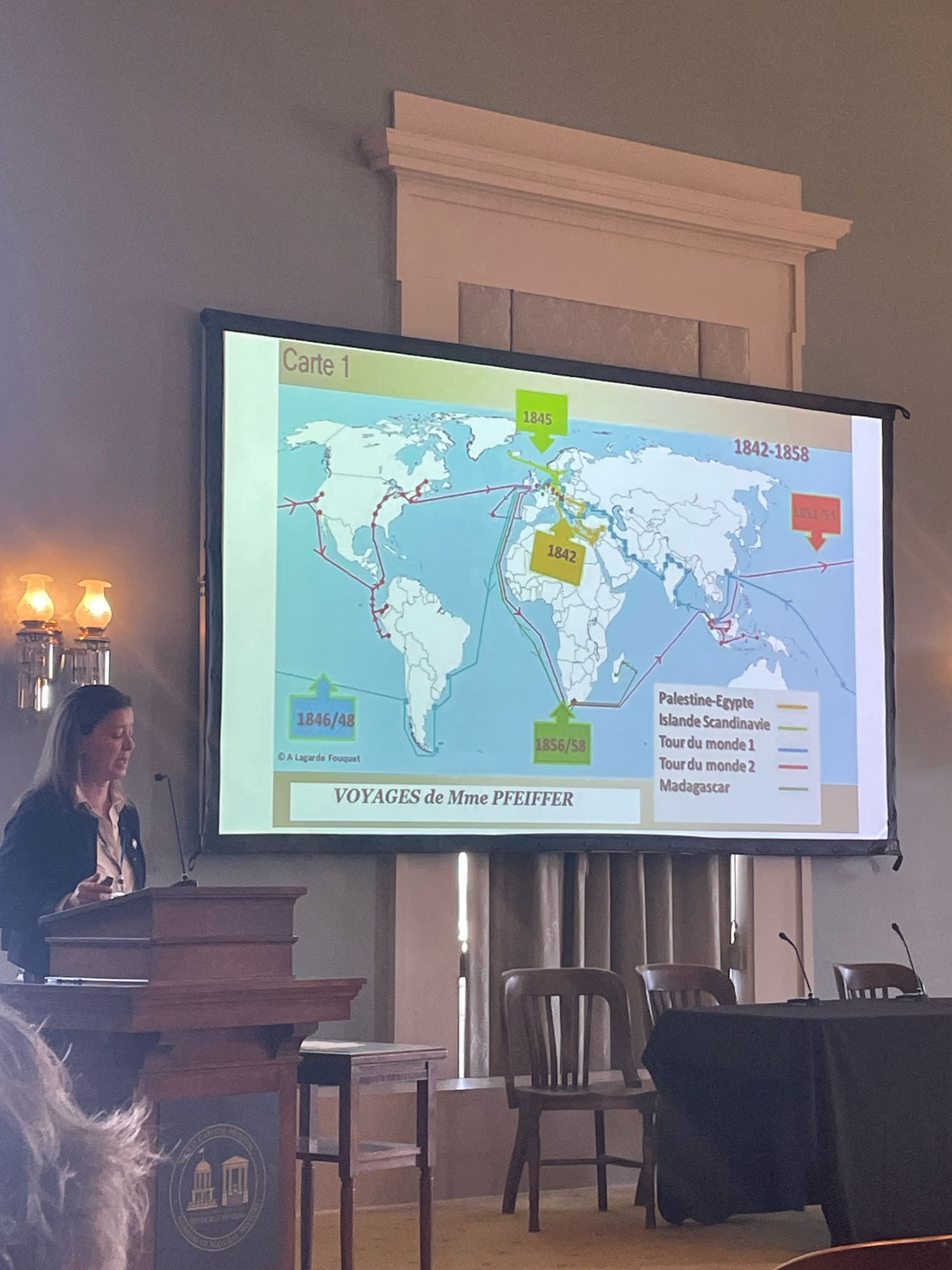 Woman stands at podium in lower left foreground. Projection screen in center shows map of world travels.