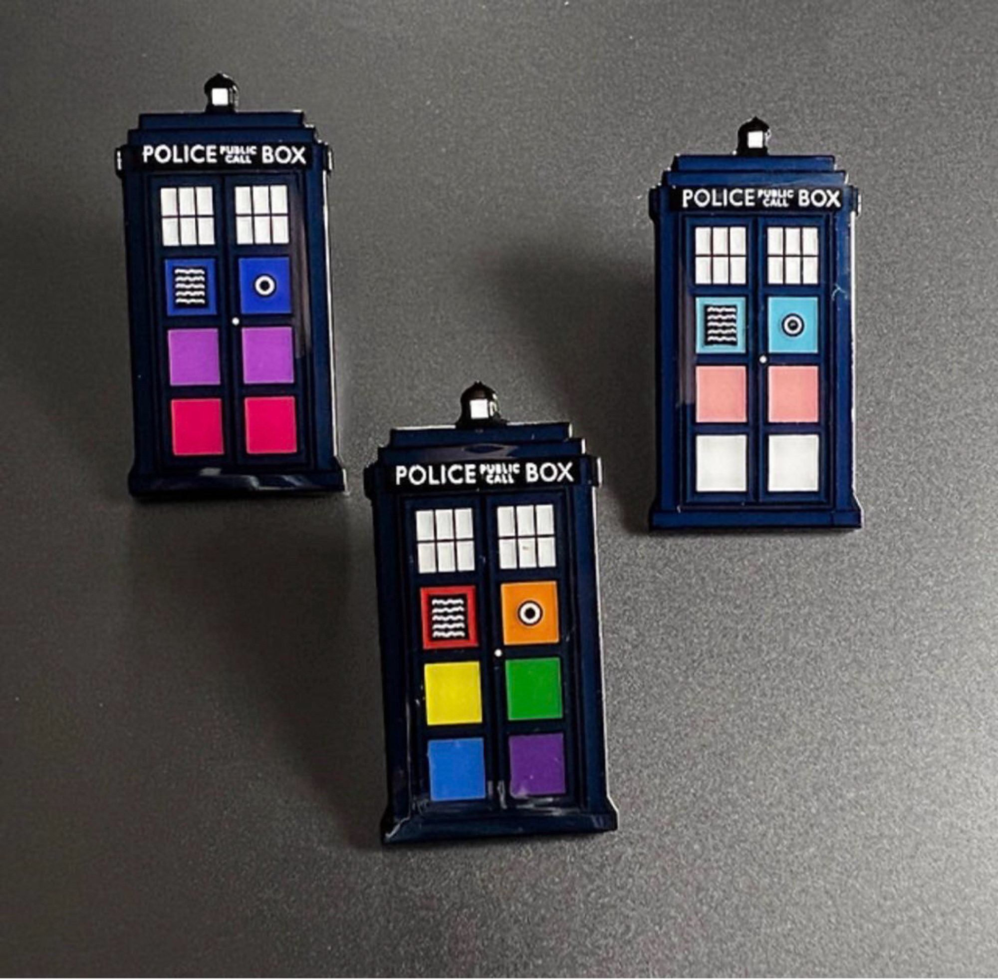 Three tardis pins. Each showing the colour of a different pride flag. Bi, rainbow and trans.