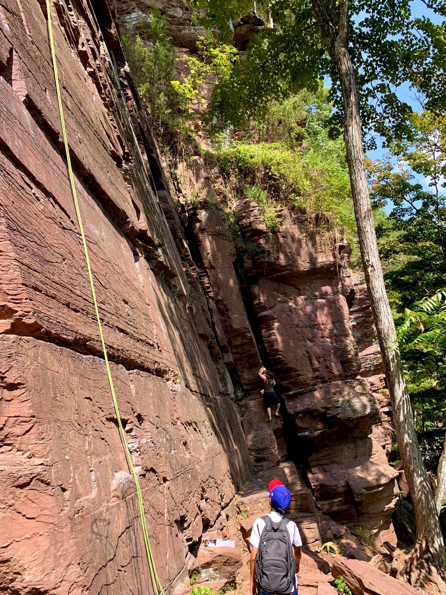 A light green rope hangs from the top of the red rock formation. A boy walks along a trail below, while a woman attempts to climb the rock without any gear.