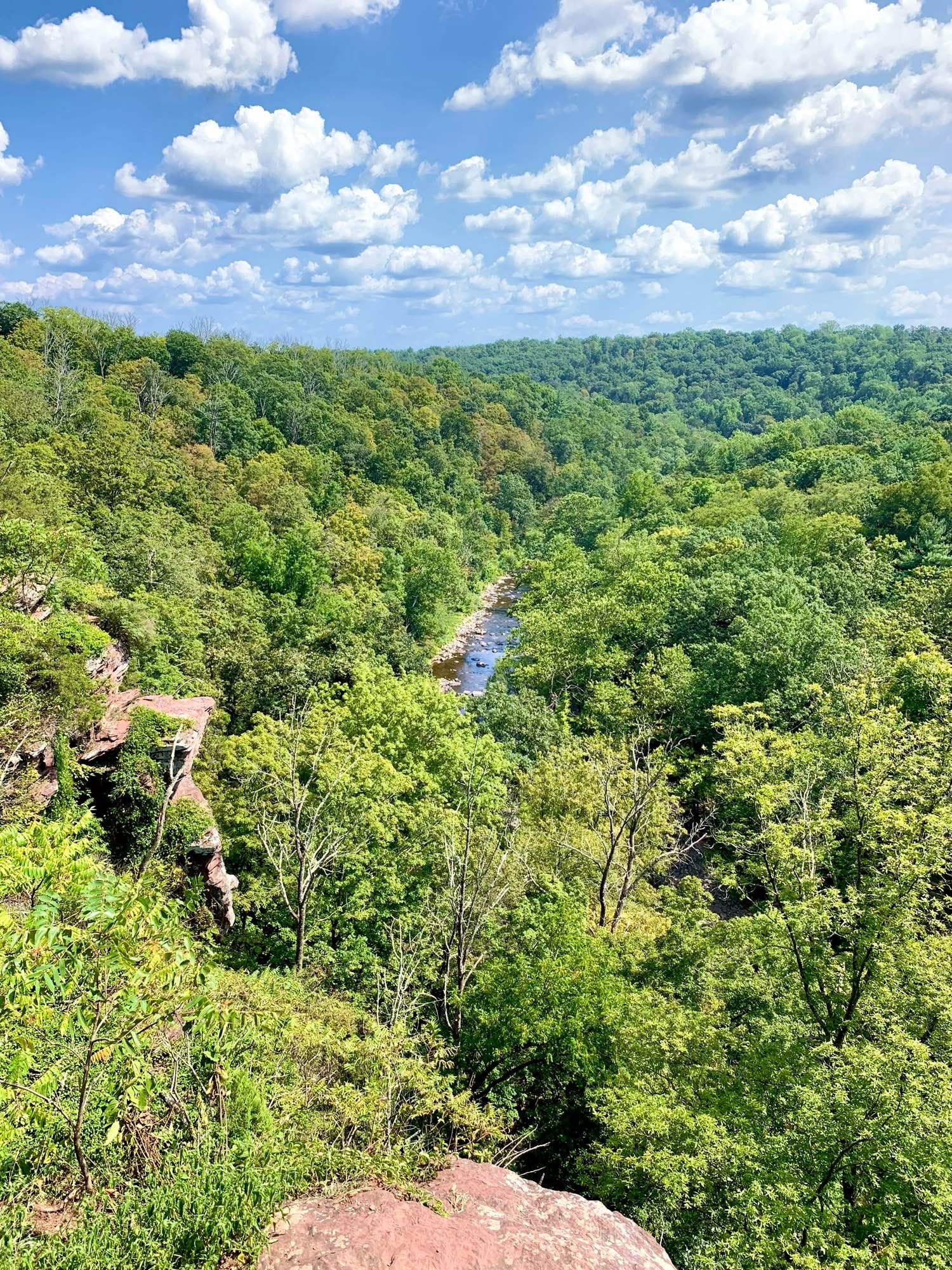 A view of Tohickon Creek flowing through the forest in the valley below. It's a sunny day with a few distinct clouds against the blue sky. Two people are sitting on a rock in the creek.