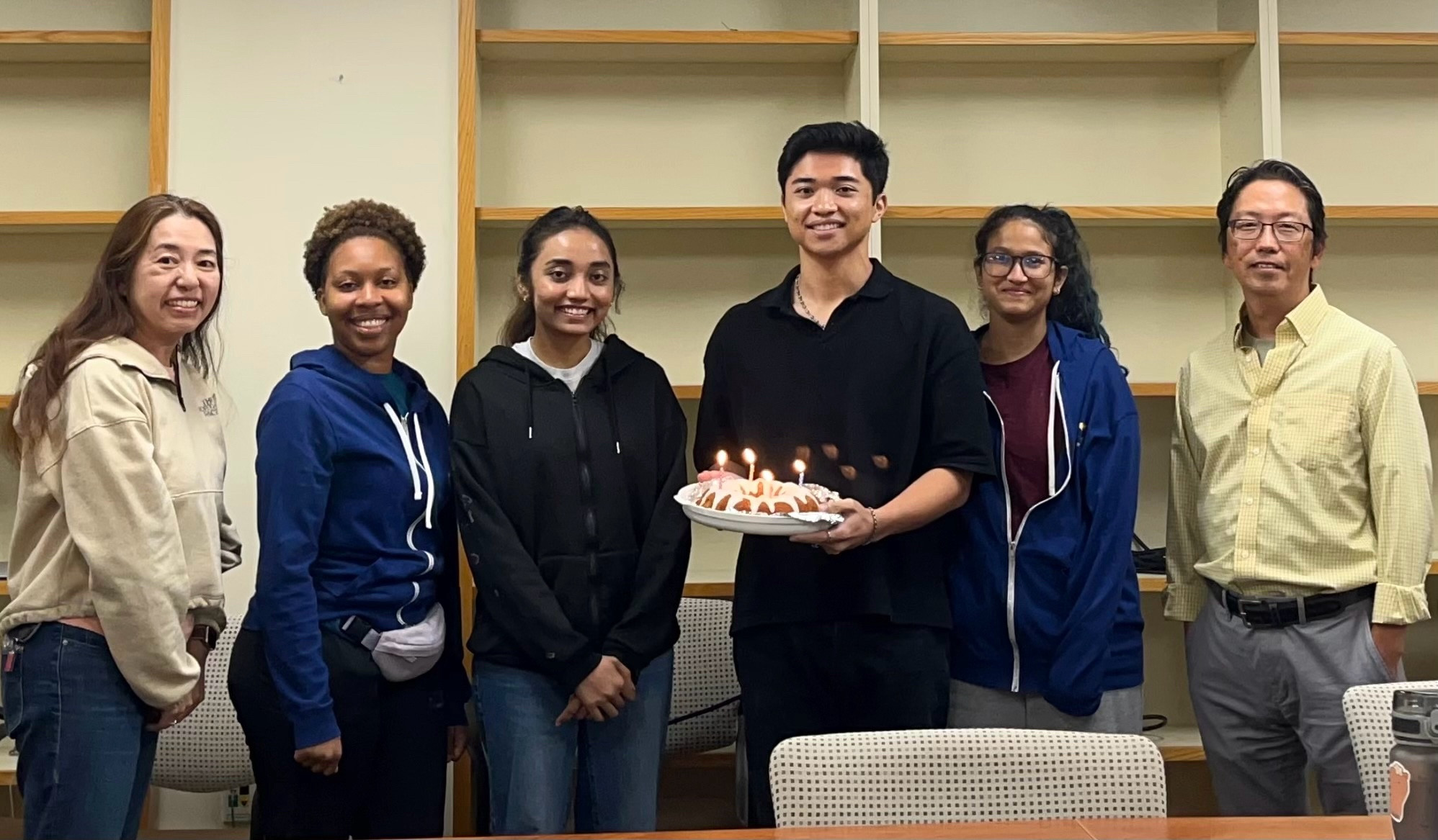 In the photo, six people are standing in front of shelves, all smiling. The tallest person is holding a birthday cake.