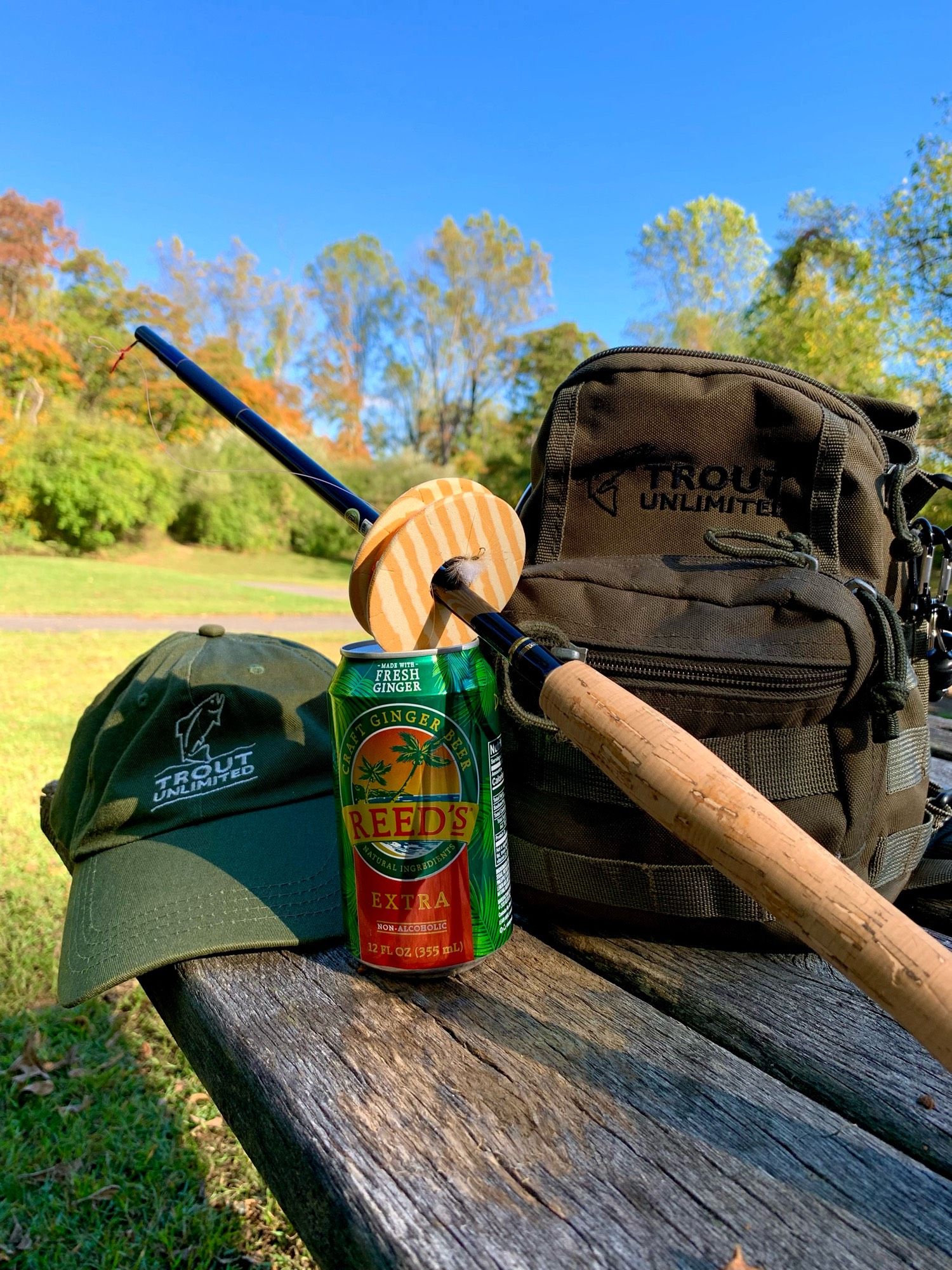 A tenkara rod, small fishing bad, a hat, and ginger beer are on a picnic table. Woods and the blue sky are in the background. The bag and hat have the trout unlimited logo.