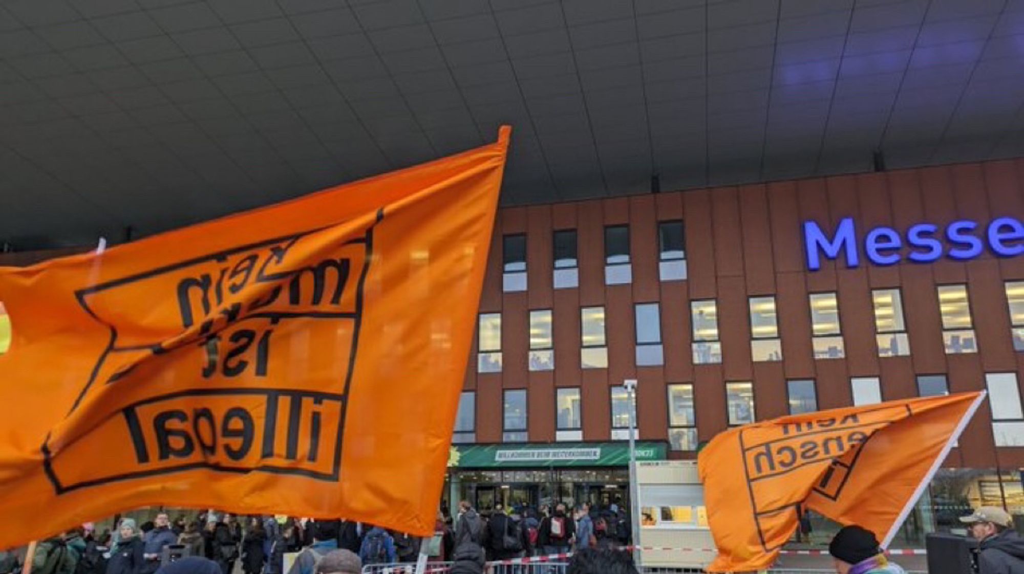 Demonstrant*innen vor der Messe Karlsruhe. Eine große „Kein Mensch ist illegal“-Flagge im Vordergrund