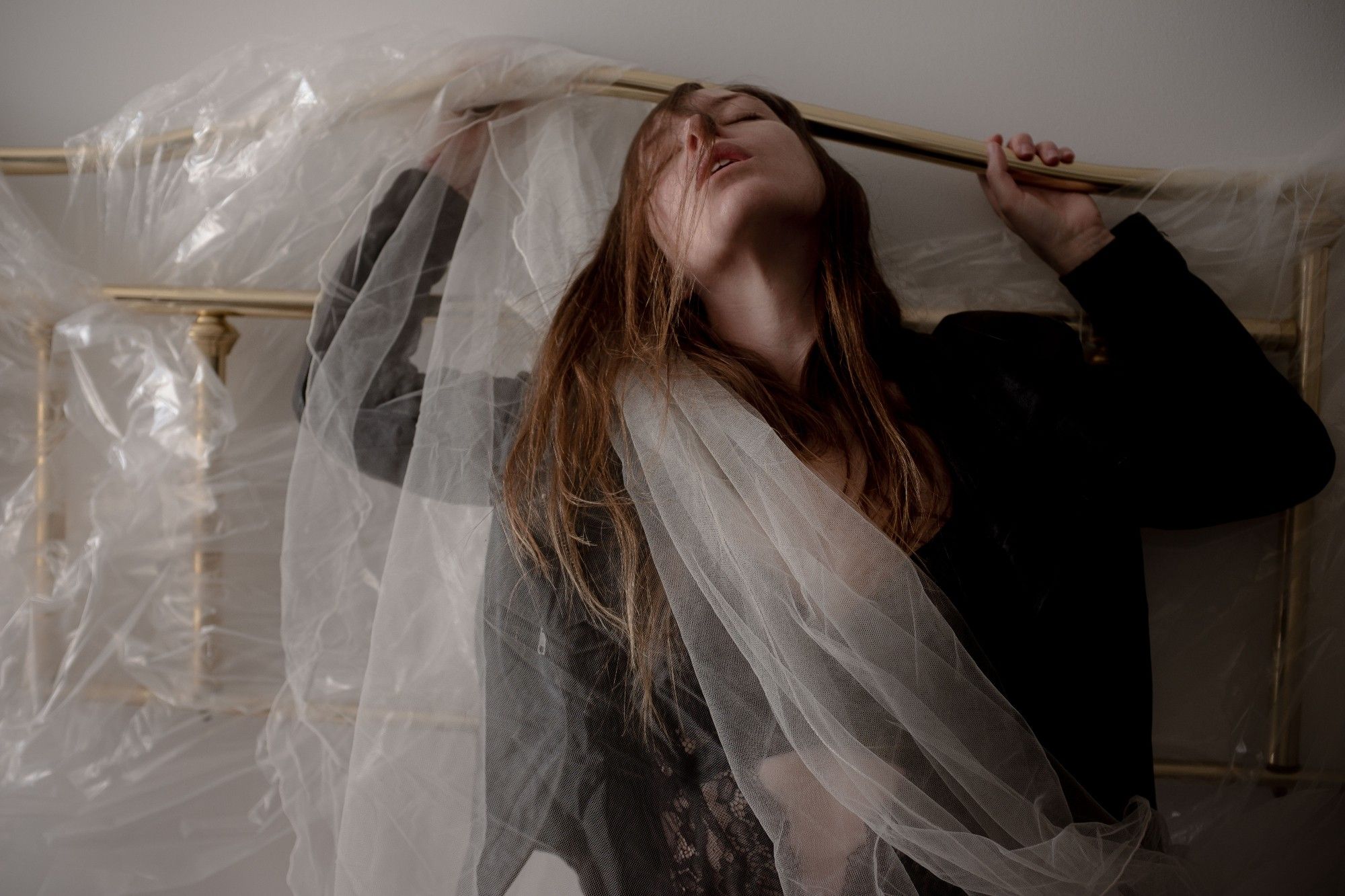 a brass headboard, disembodied from a bed pressed against the wall draped in clear shiny plastic and pale white tulle. A person grips the headboard rail, head tossed back so their hair falls across their face. they are wearing a leather motorcycle jacket and a criss cross scrap of black lace