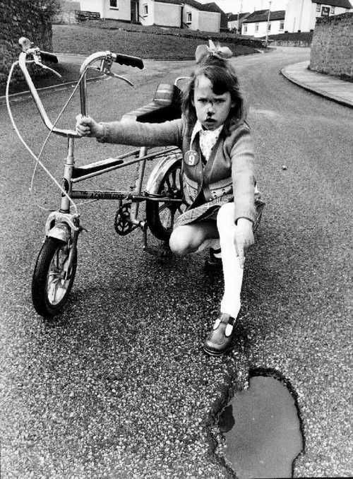 Amanda Groves, aged nine, injured when her bike hit a pothole in Escomb, Co. Durham (1972, Northern Echo)

Small girl with a bloodied nose and an “I am 9” badge, points wanly at a water-filled pothole. Her bike is a Raleigh Tomahawk, the junior version of the Chopper. 