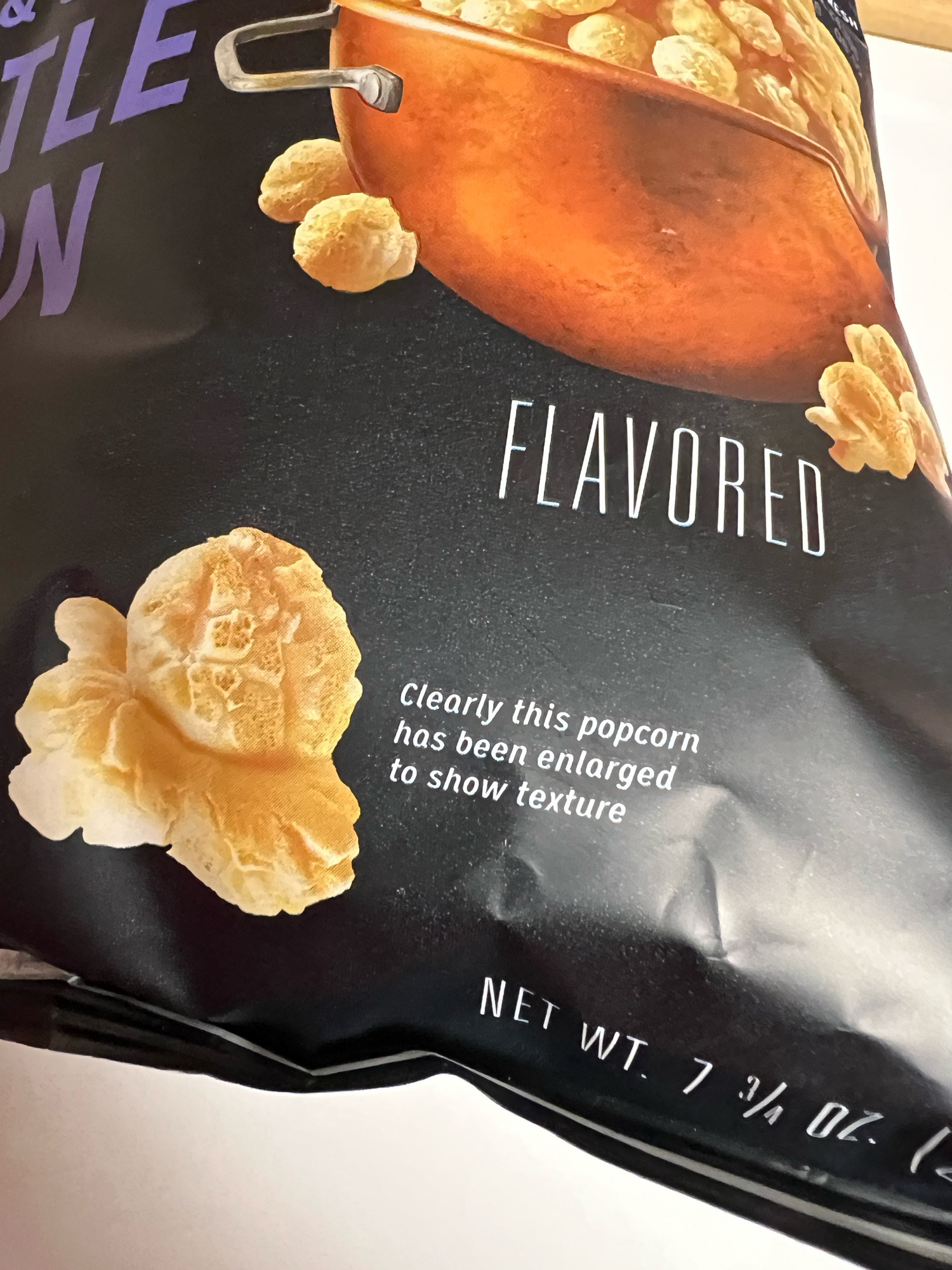 Photo of the bottom half of a bag of Smartfood kettle corn with an image of a popped kernel and the words “Clearly this popcorn has been enlarged to show texture” next to it. 