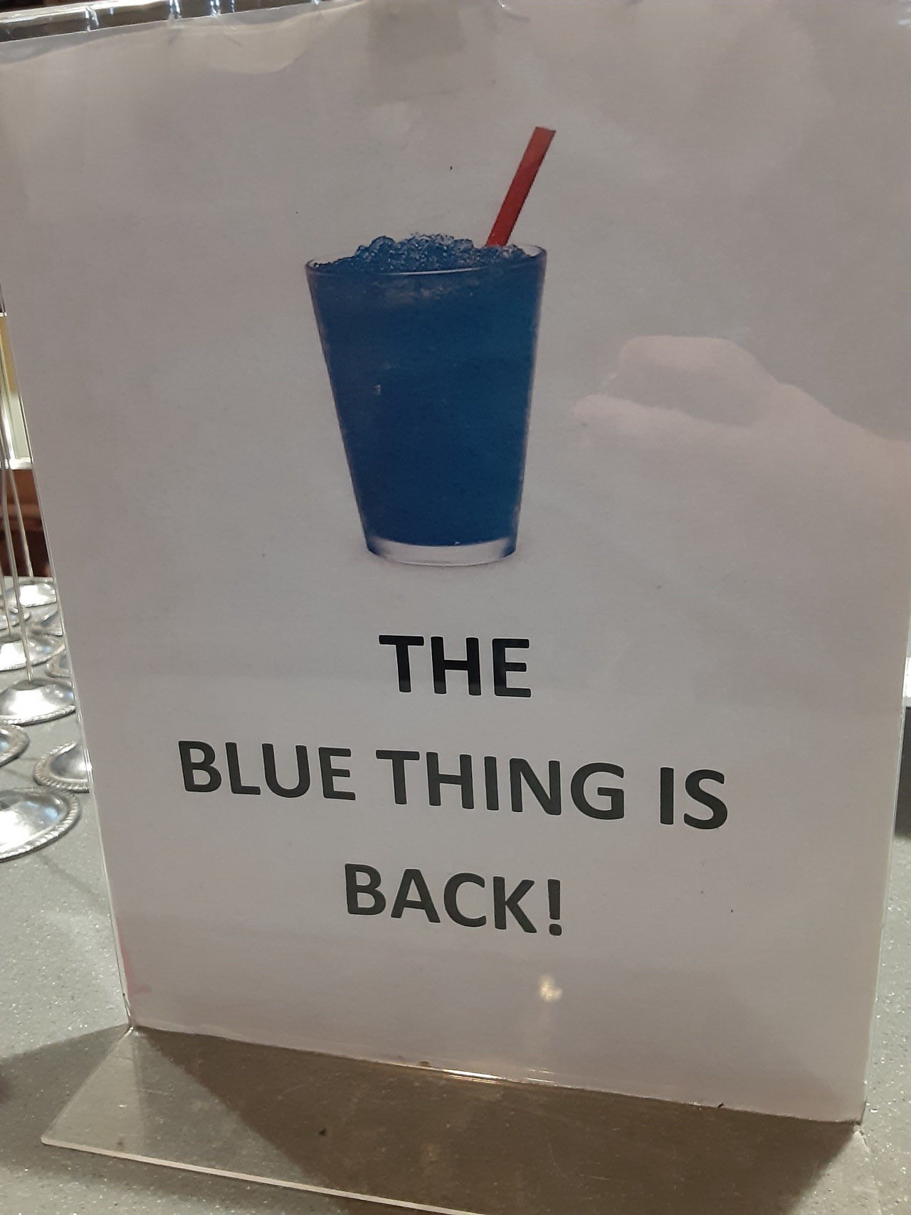 Photo of a crudely printed paper sign at a restaurant. The sign has a stock photo of a blue slush drink. “The blue thing is back!”, is printed below in all caps.