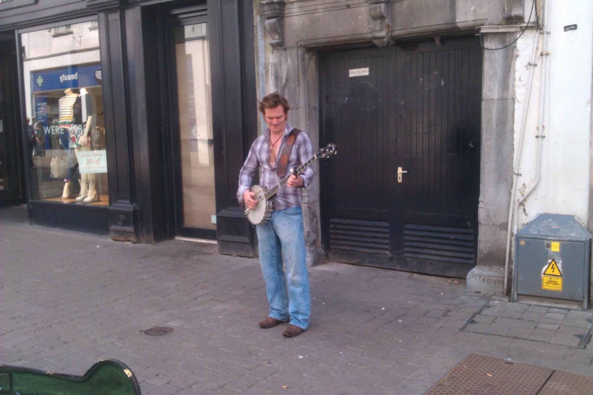 Vor einem Garagentor steht ein junger Mann in Jeans und offenem Karohemd mit einem Banjo umgehängt.