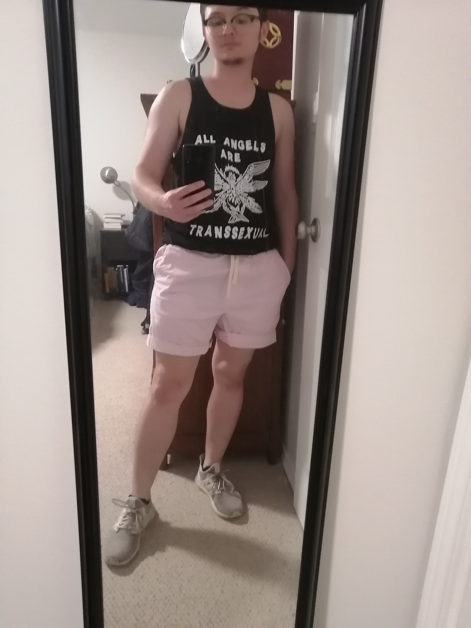 Mirror selfie of me wearing a black tank top with 'all angels are transsexual' printed on it in white with an abstract drawing of an angel (all wings and eyes) in the center. Also light pink shorts and white running shoes