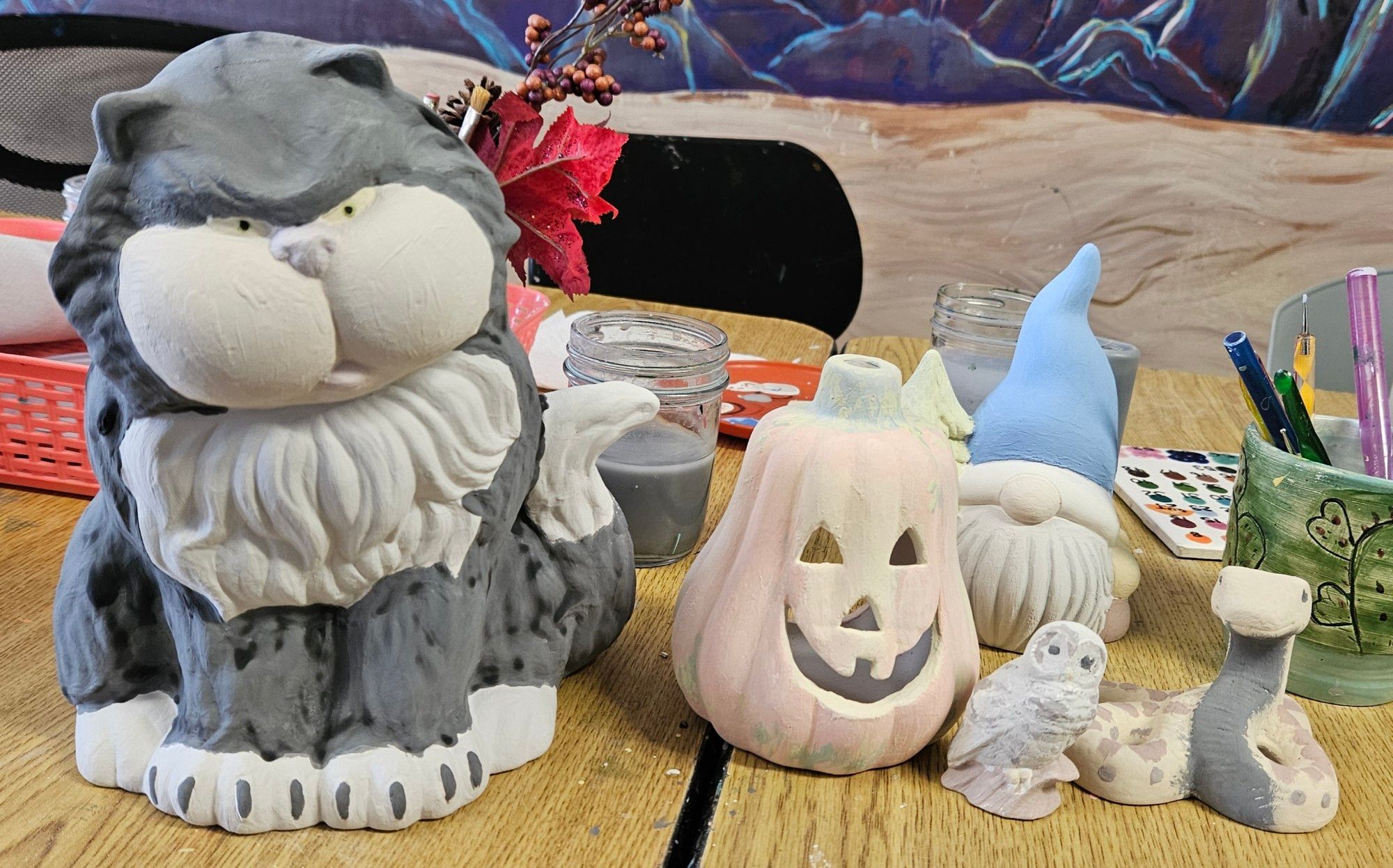 Ceramic statues of a large black and white fluffy cat, a jack o' lantern, a winter gnome, a barn owl, and a snake. Painted in muted glaze colors that will change after time in the kiln. On a table with painting supplies in the background.