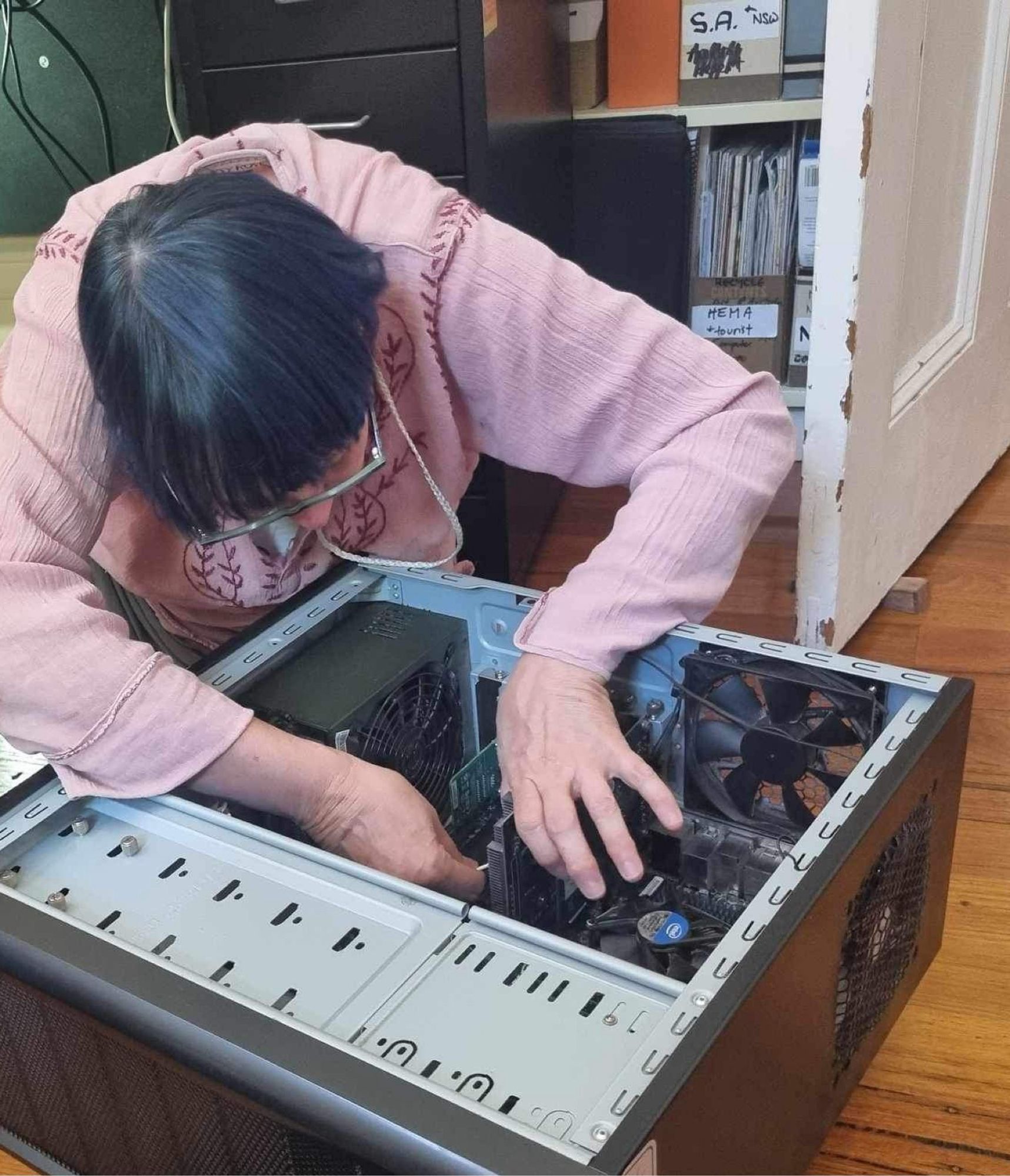 photo, computer tower lying on the floor with a side panel removed, I'm bent over peering into it and reaching inside it to do repair