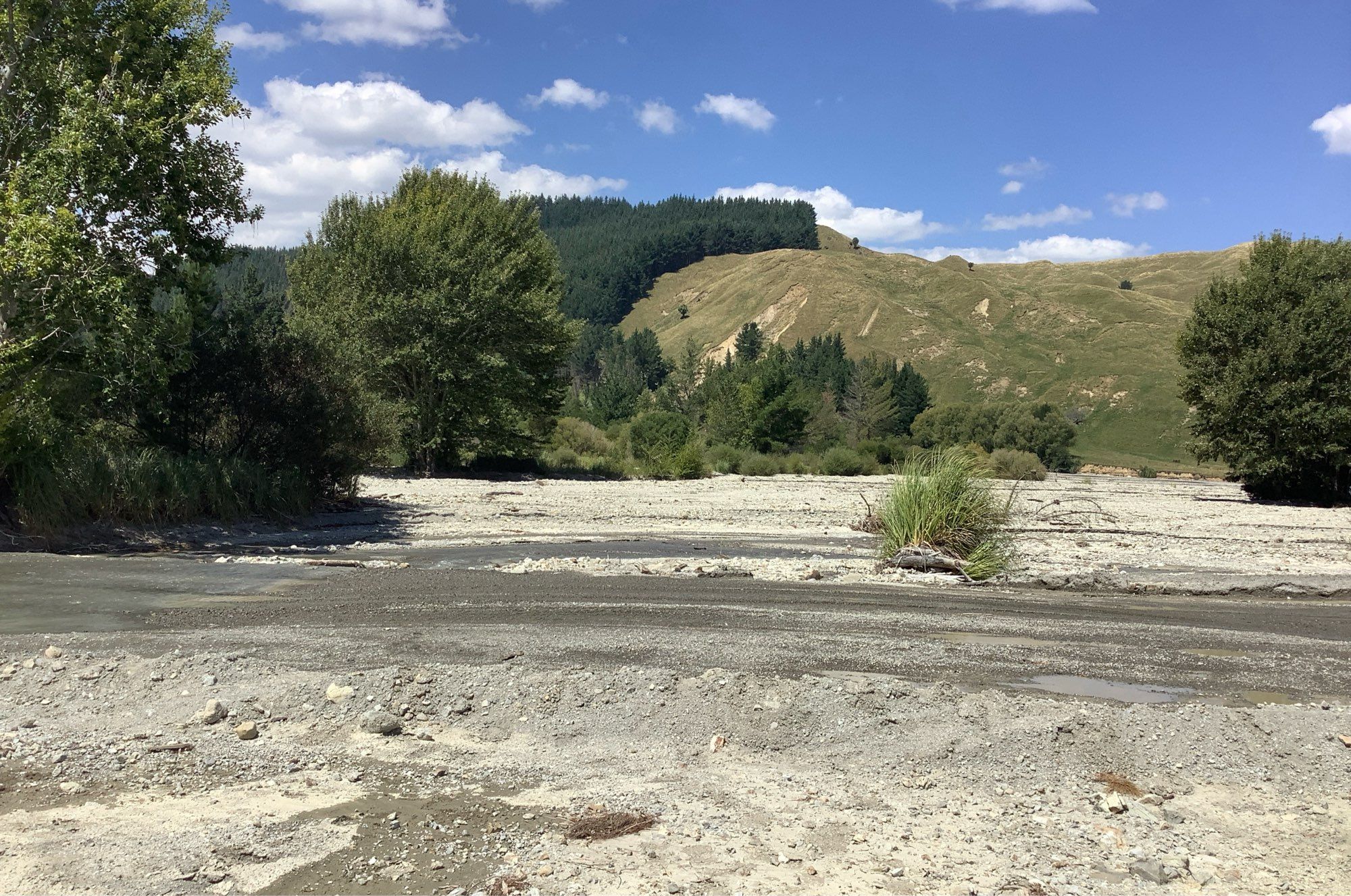 view across a river bed that's choked with coarse and very poorly sorted sediment, hills on the opposite side of the valley show pale scars of recent landsides