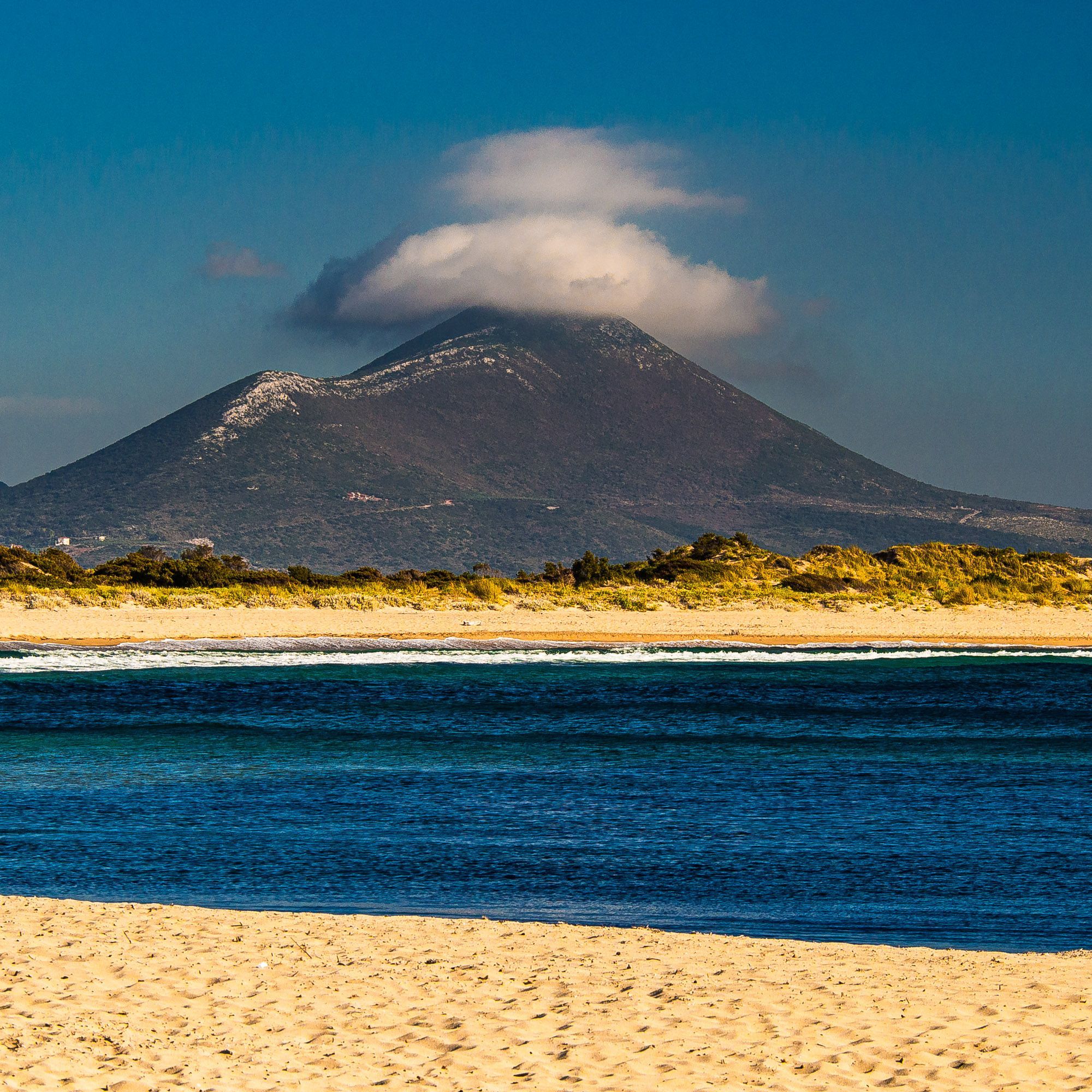 Es ragt ein Berg in die Höhe, auf der Spitze des Berges befindet sich im ansonsten fast wolkenlosen Himmel eine dicke Wolke. Der Berg ist weit nach hinten. Davor sieht man einen Strand mit Dünen, davor das dunkelblaue Meer und wieder davor noch mal Strand.