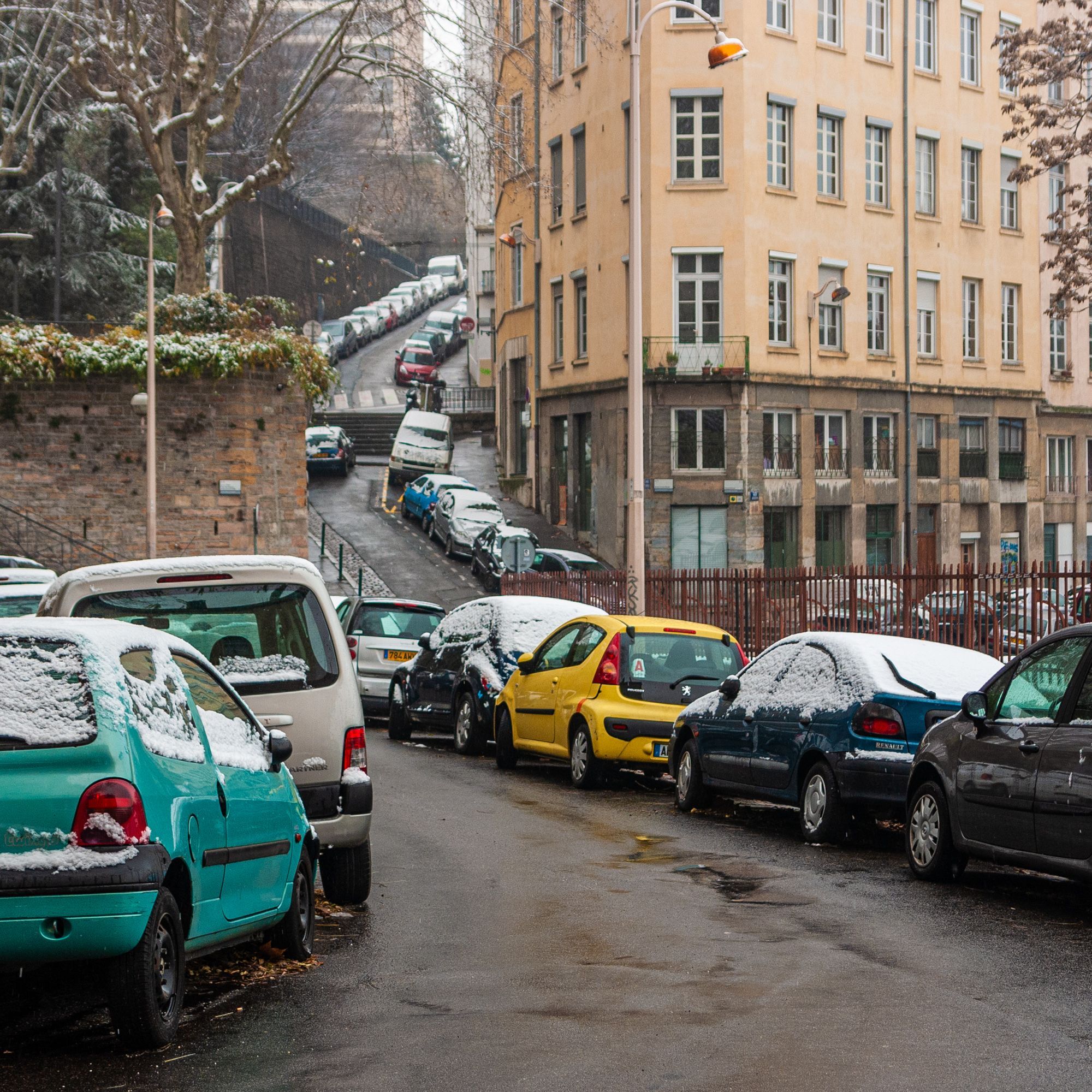 Eine grau-nasse Straße die im Vordergrund abschüssig ist und danach in einer Kurve einen Berg hochführt. Links und rechts stehen Autos geparkt mit einer dünnen Schneeschicht. Es ist etwas neblig-trüb und die Szene wirkt trotz bunter Autos etwas farblos.