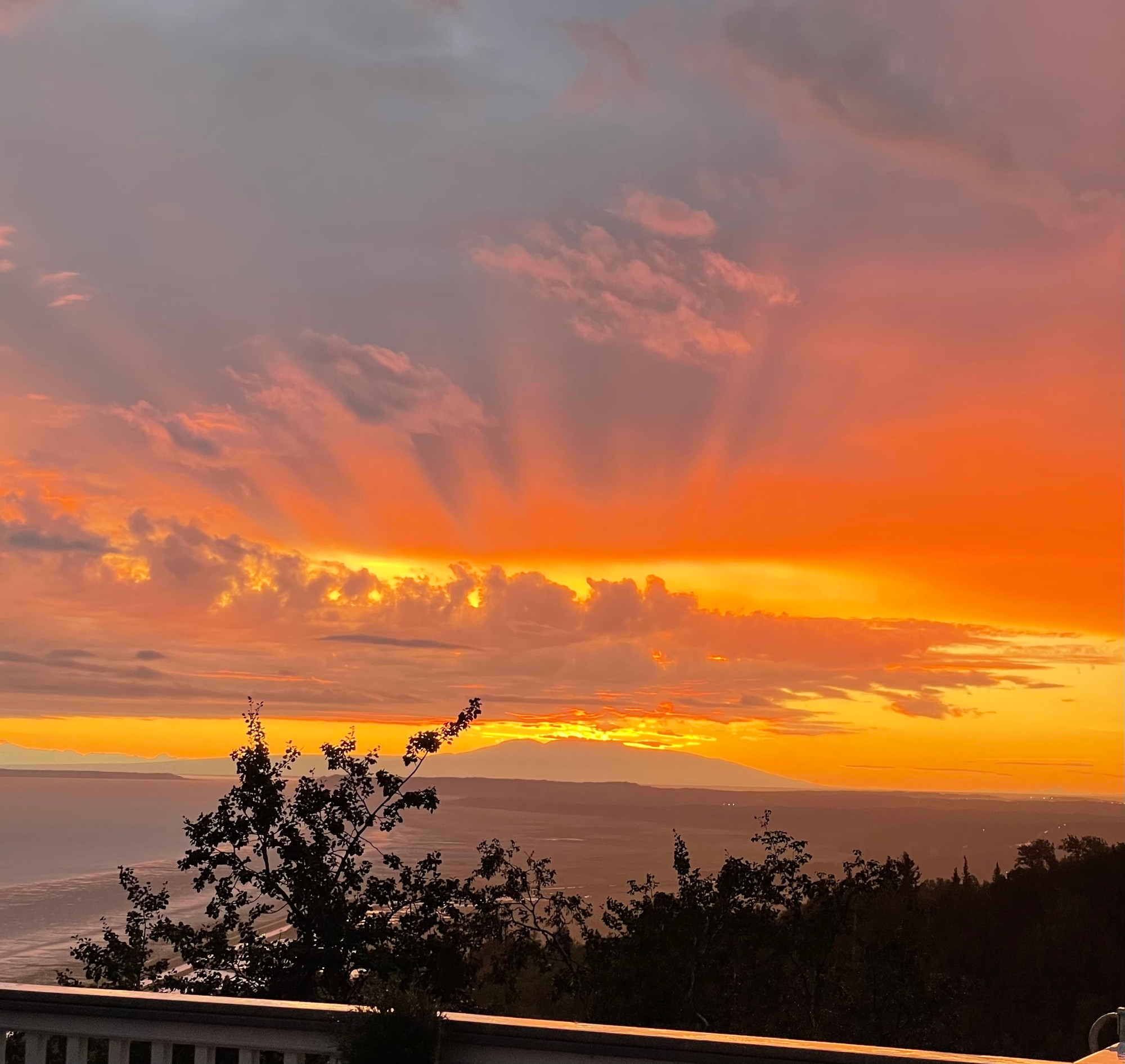 A balcony view of Cook Inlet, with Sleeping Lady (Mount Susitna) in the distance. There are trees in the foreground, and the water is looking a lilac color. The sky is vibrant with the setting sun casting yellow and orange upon the clouds. 

Anchorage, AK 
August 2025