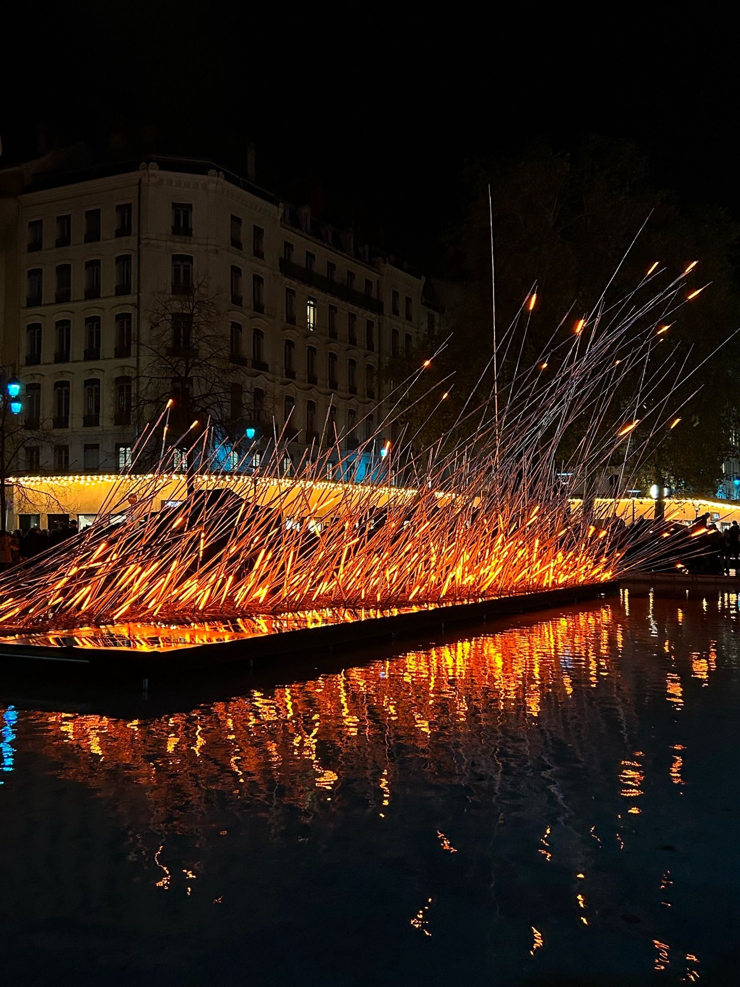 Photo d’une installation de la fête des Lumières de Lyon 2023, place de la République. Des fils métalliques illuminé en orange à la base donnant l’illusion d’un feu.