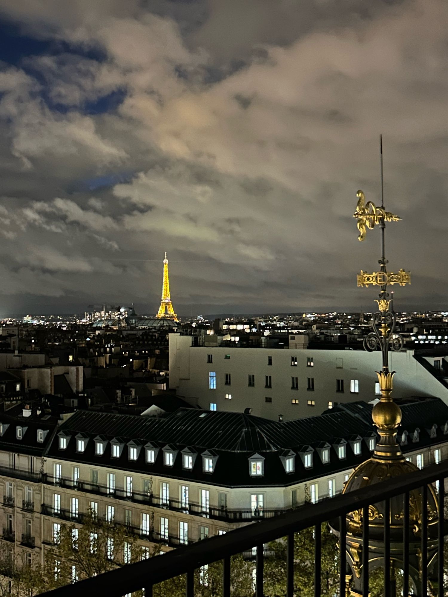 Vue de Paris de nuit depuis le toit du Printemps Haussmann, avec la Tour Eiffel illuminé à l’arrière plan et la flèche du magasin avec une girouette au premier plan.