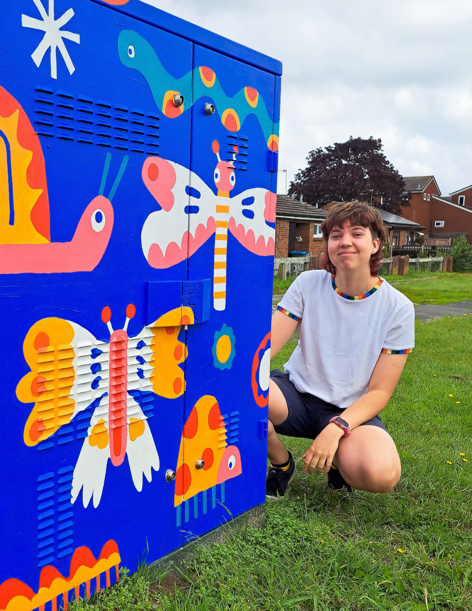 A photo of me crouched next to an electrical box mural I painted. The design is full of colourful bugs and butterflies on a bright blue background. 