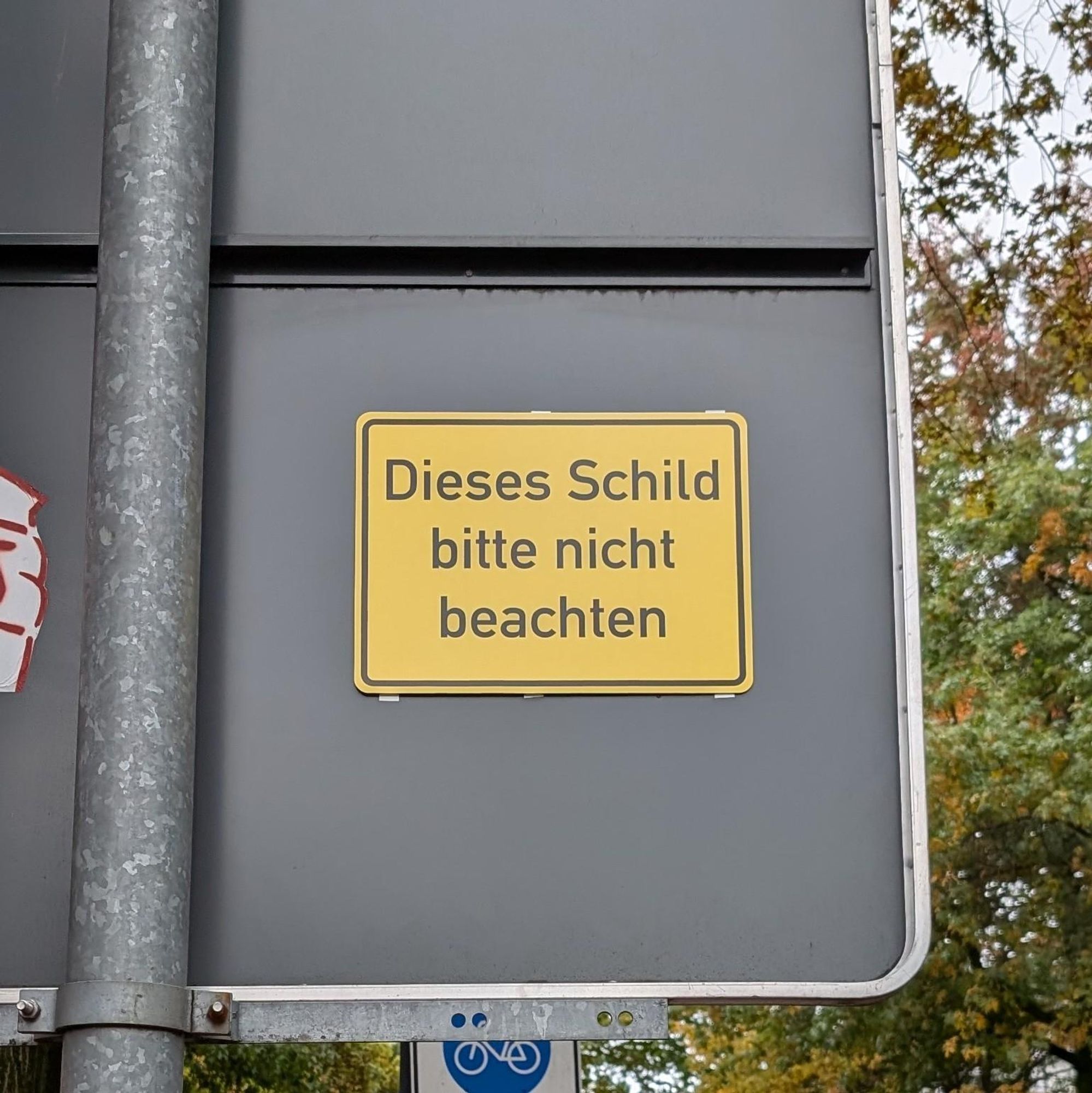 Gelbes Schild in Optik eines Ortseingangsschilds hinten auf ein reguläres Verkehrsschild geklebt. Beschriftung:

"Dieses Schild bitte nicht beachten"