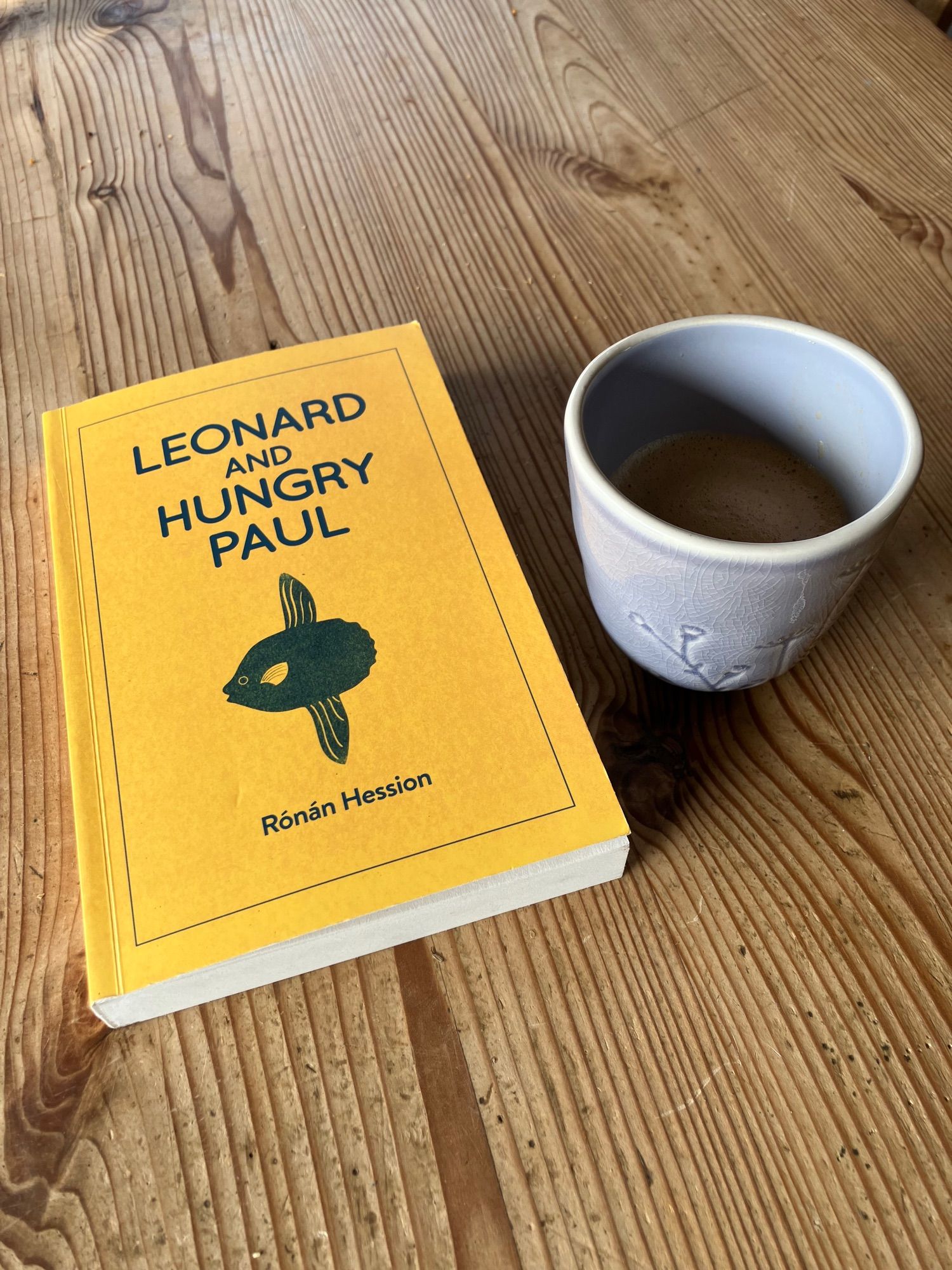 Leonard and Hungry Paul by Rónán Hession on a wooden table. A handless lilac cup containing coffee sits alongside.