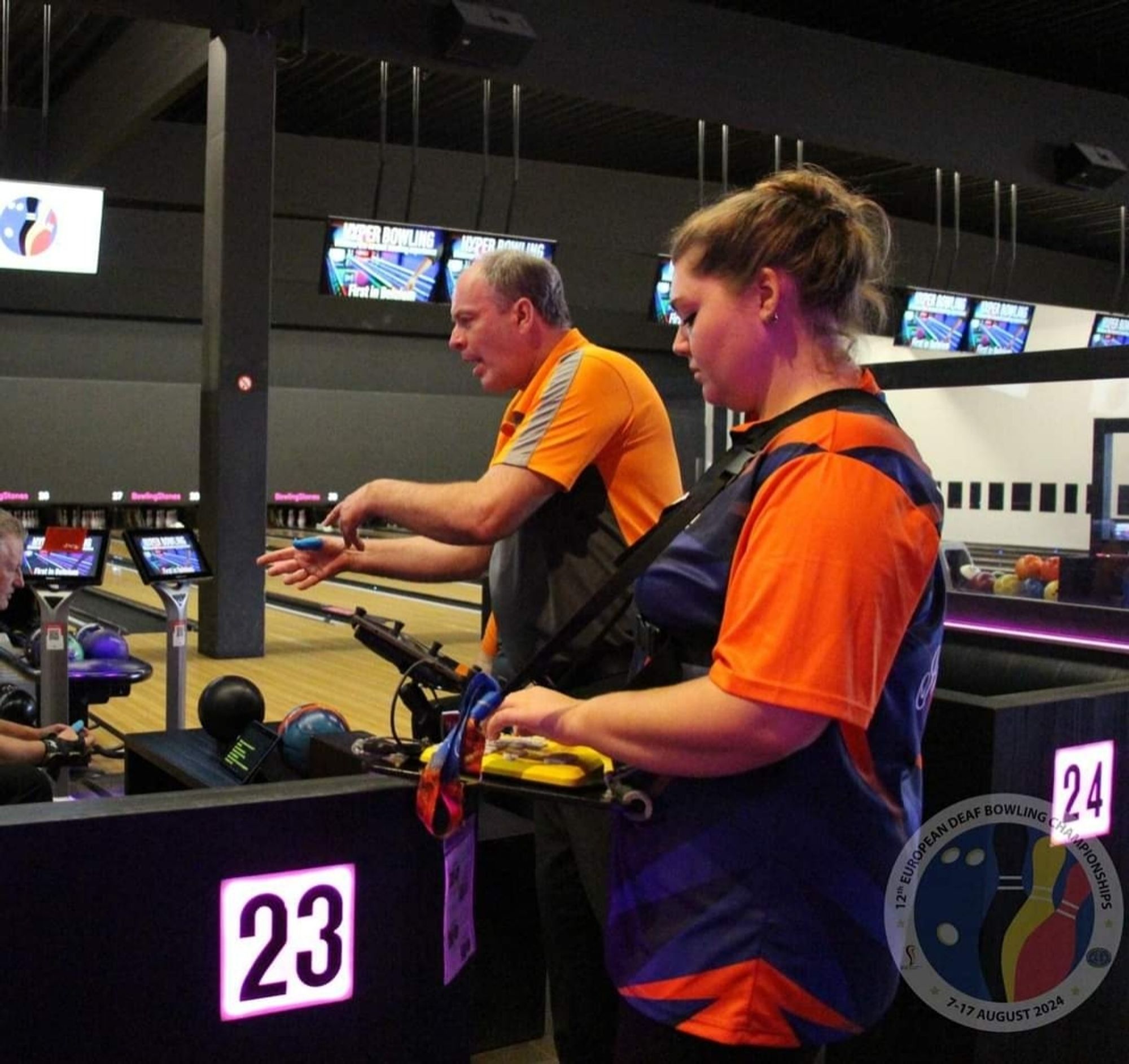 Gekleed in een oranje  shirt met korte mouwen staat Roselinde met opgestoken bruin haar naast een mannelijke deelnemer bij baan 23. Ze heeft op haar velowalker een geel toetsenbord liggen waarmee ze haar tolktekst in beeld brengt voor iedere dove/sh deelnemer