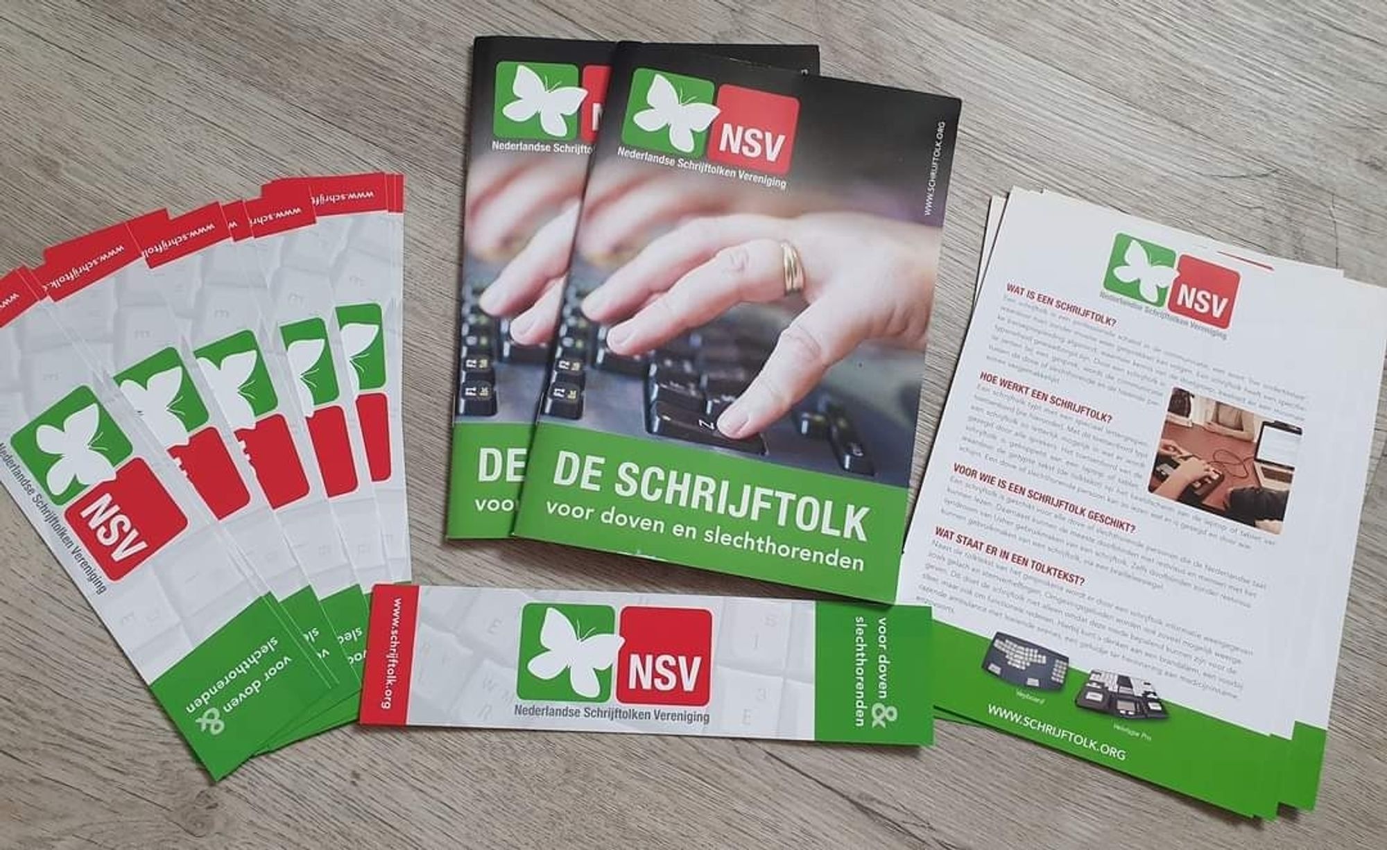 Op een grijze tafel liggen promotiematerialen
Links in waaiervorm een 6 boekenleggers met de kleuren groen en rood. Op een groene achtergrond een witte vlinder en op een rode achtergrond met witte letters: NSV
In het midden liggen 2 brochures met een foto van de hand van een schrijftolk boven haar toetsenbord.  En onderaan in groene balk de titel: de schrijftolk. 
Aan de rechterkant liggen gestapeld een 3 flyers met oa logo en foto van de 2 meest gebruikte toetsenborden voor schrijftolken. Velotype pro en veyboard