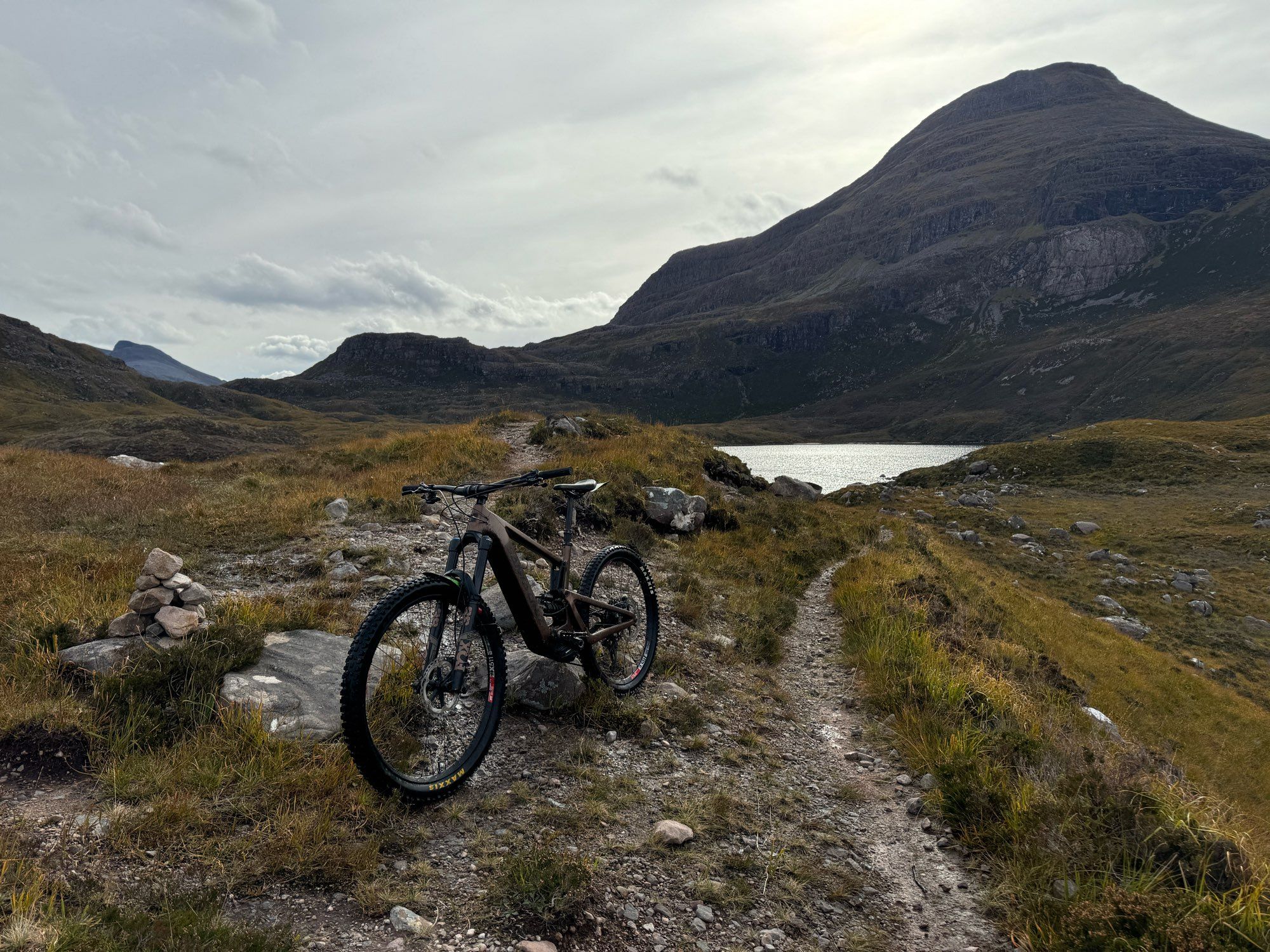 Scottish highlands.  A mountain bike in the foreground, facing toward the camera. A rough, single track path goes off into the distance on the right, towards a hill loch that can just be glimpsed. In the background, a steep sided Munro, Maol Chean-dearg. High clouds in the sky.