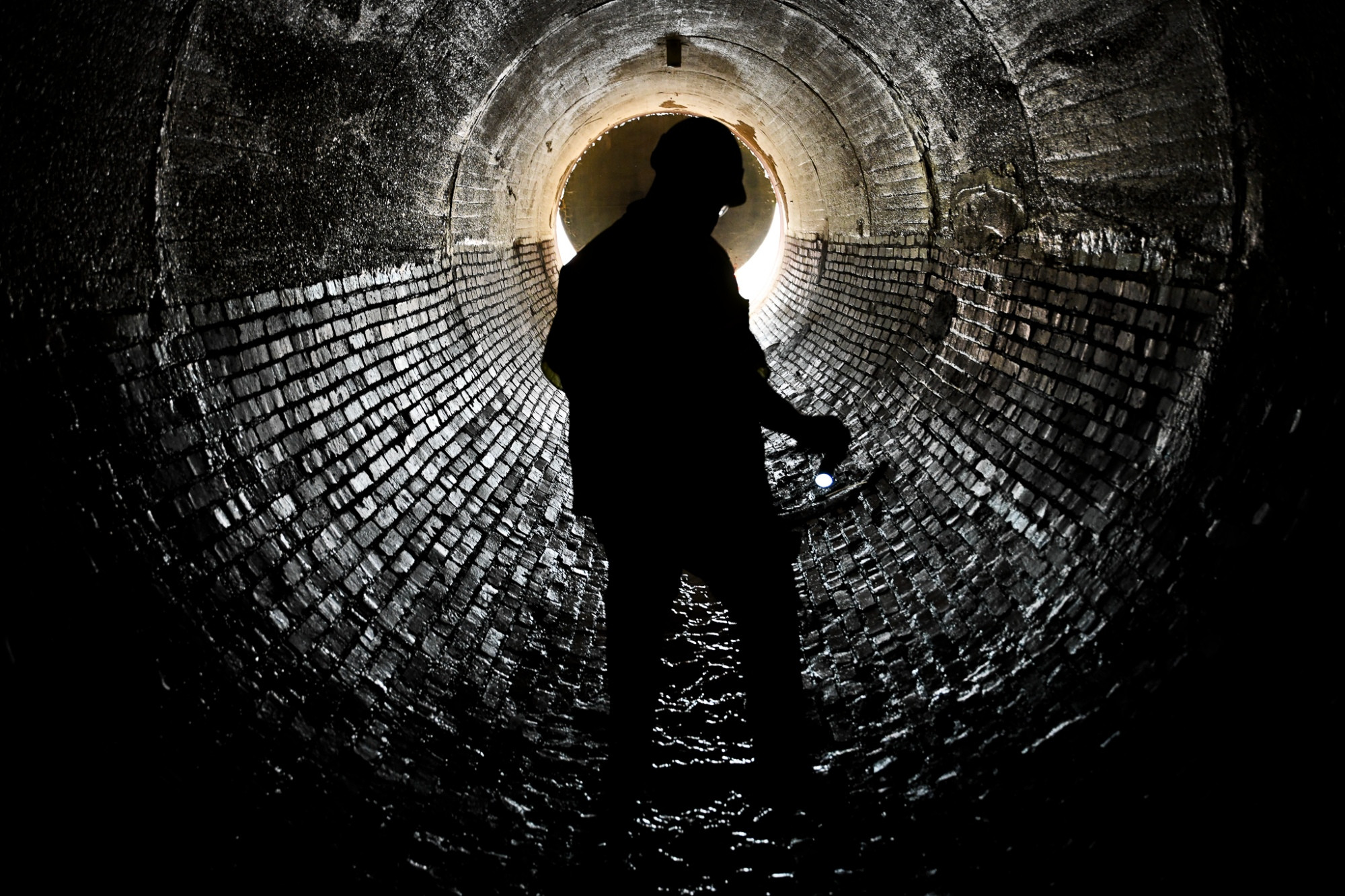 a silhouetted figure stands in a long brick sewer tunnel illuminated from afar where the tunnel ends. the brick work reflects the distant light and the person in shadows looks downward to ensure they are stepping safely.