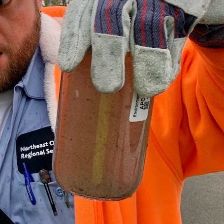 a gloved hand holds a jar of cloudy brown sewage.