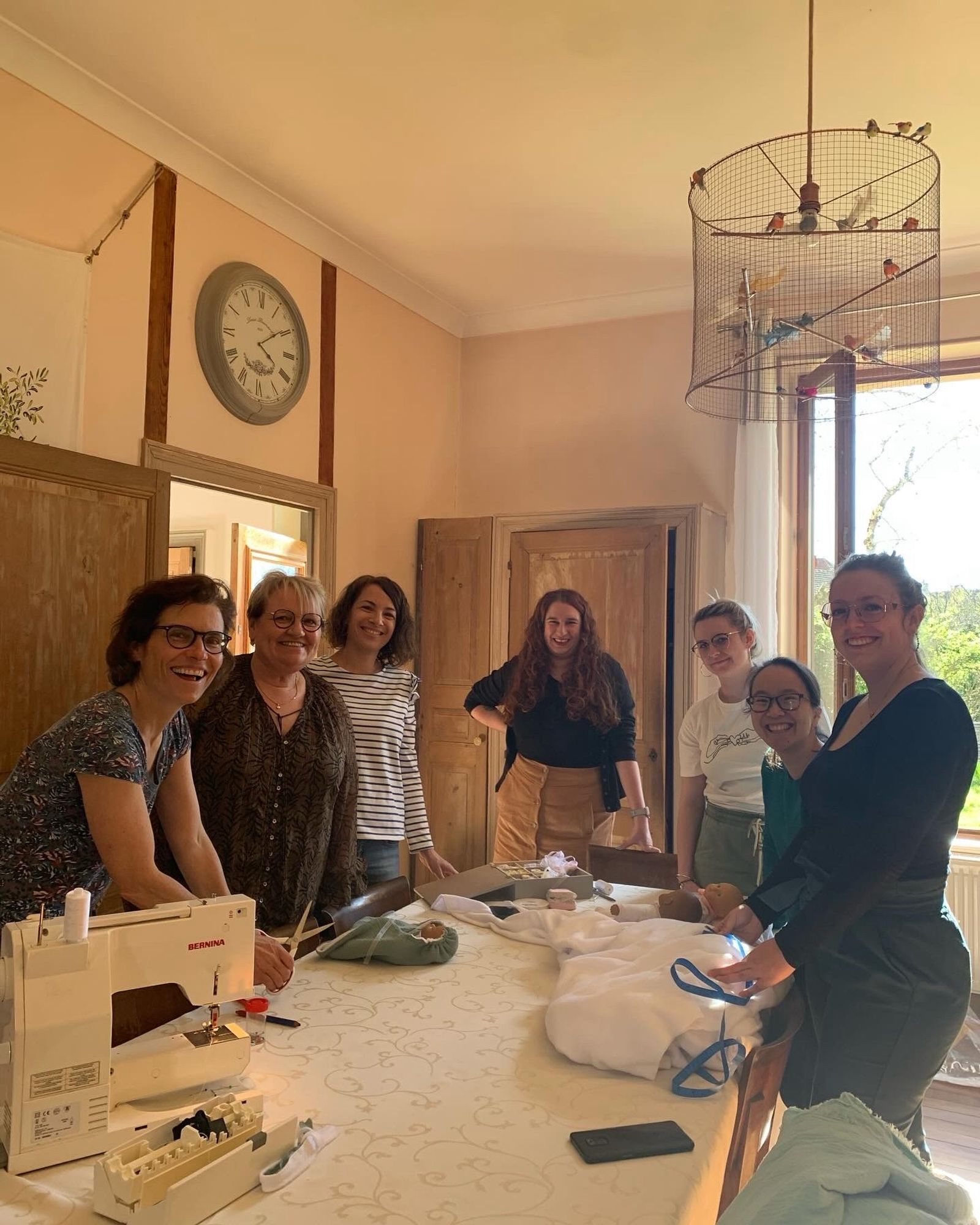 7 femmes autour d’une table sur laquelle se trouvent une machine à coudre, du tissu déroulé, un patron de nid d’ange