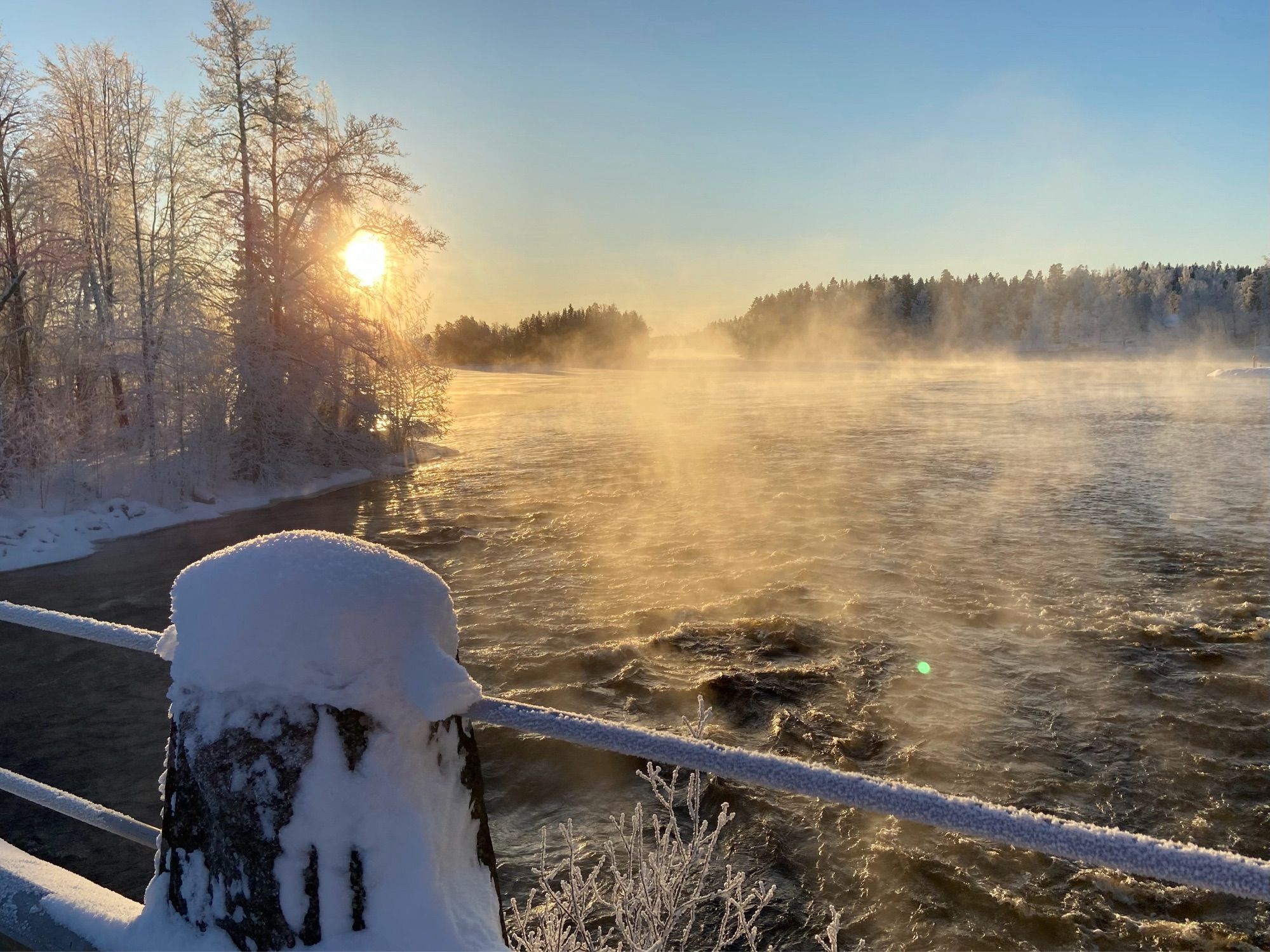 Vesihöyry nousee koskesta aurinkoa vasten muodostaen hienoja utukuvia kuin henkihahmoja. Taivas on pilvetön ja niin sininen.