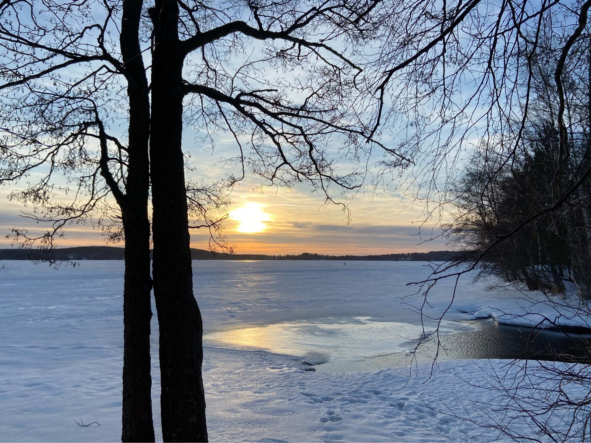 Aamun aurinko nousee järven takaa keltaoranssina. Lammesta järveen laskeva salmi virtaa vapaana. Muutenkin jäällä näkyy vettä.