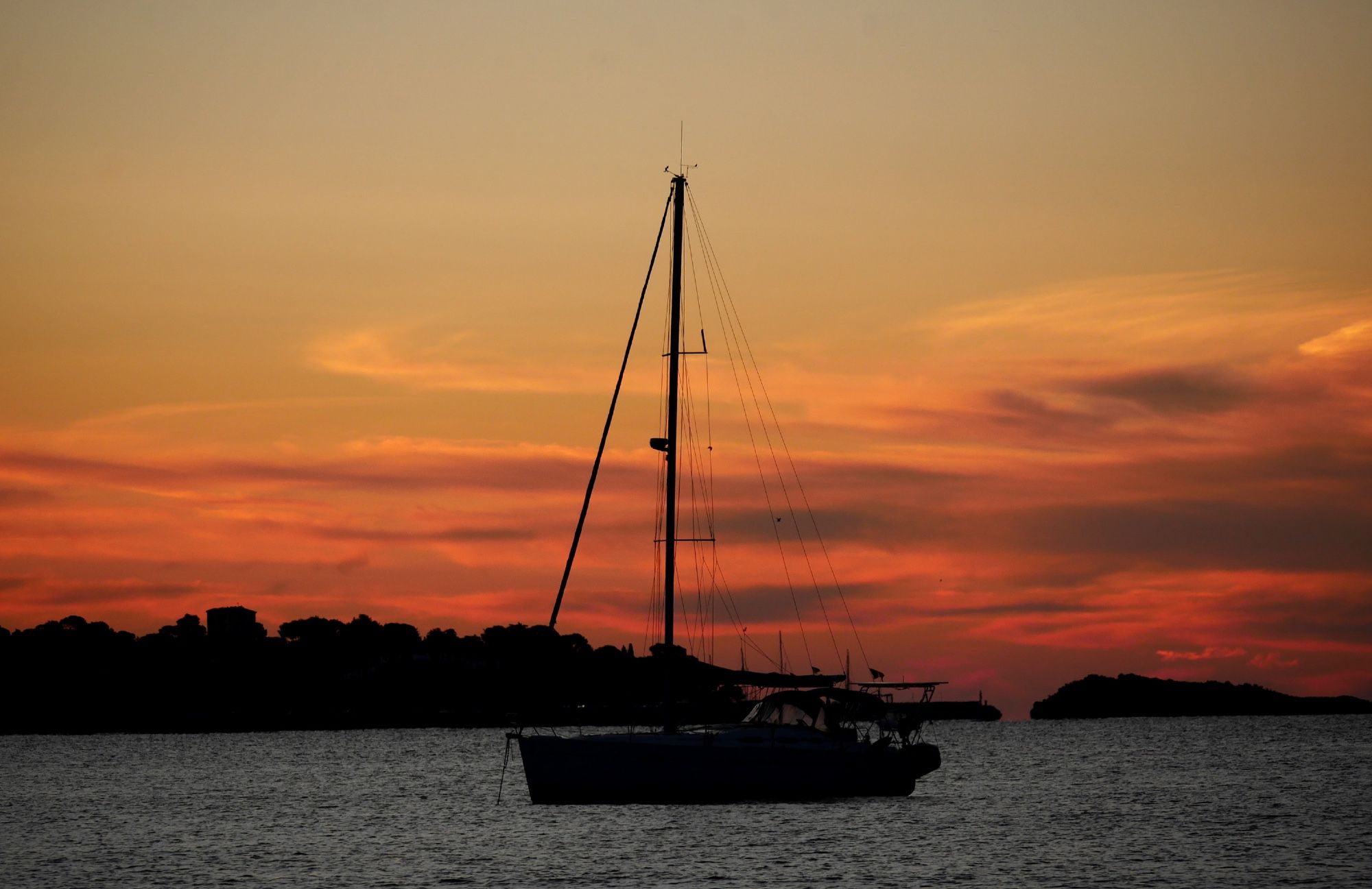 Photo d’un voilier à un mat, posé sur une mer calme couleur gris acier. Derrière le bateau une bande de terre se détache en noir sur fond de nuages colorés d’un dégradé de jaune, orange, rose et gris foncé.