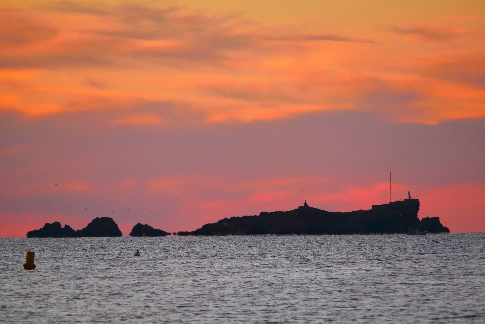 Photo d’un petit ilot de rochers dans la faible lumière de l’aurore. La mer au premier plan a une couleur gris argent. L’ilot se détache en noir sur fond de nuages colorés de rose, gris-mauve et jaune orangé.