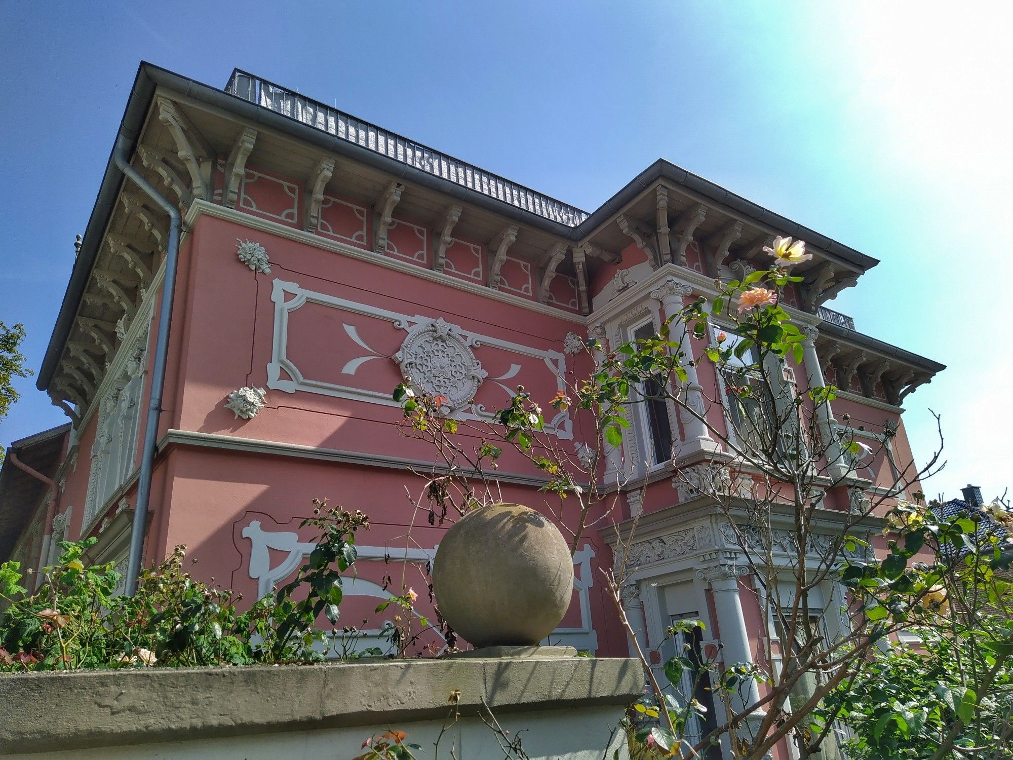 Alte Villa in rosé mit stuckverzierungen, Erkern, Figuren, davor ein blühender rosenbusch, eine Kugel auf einer Mauer