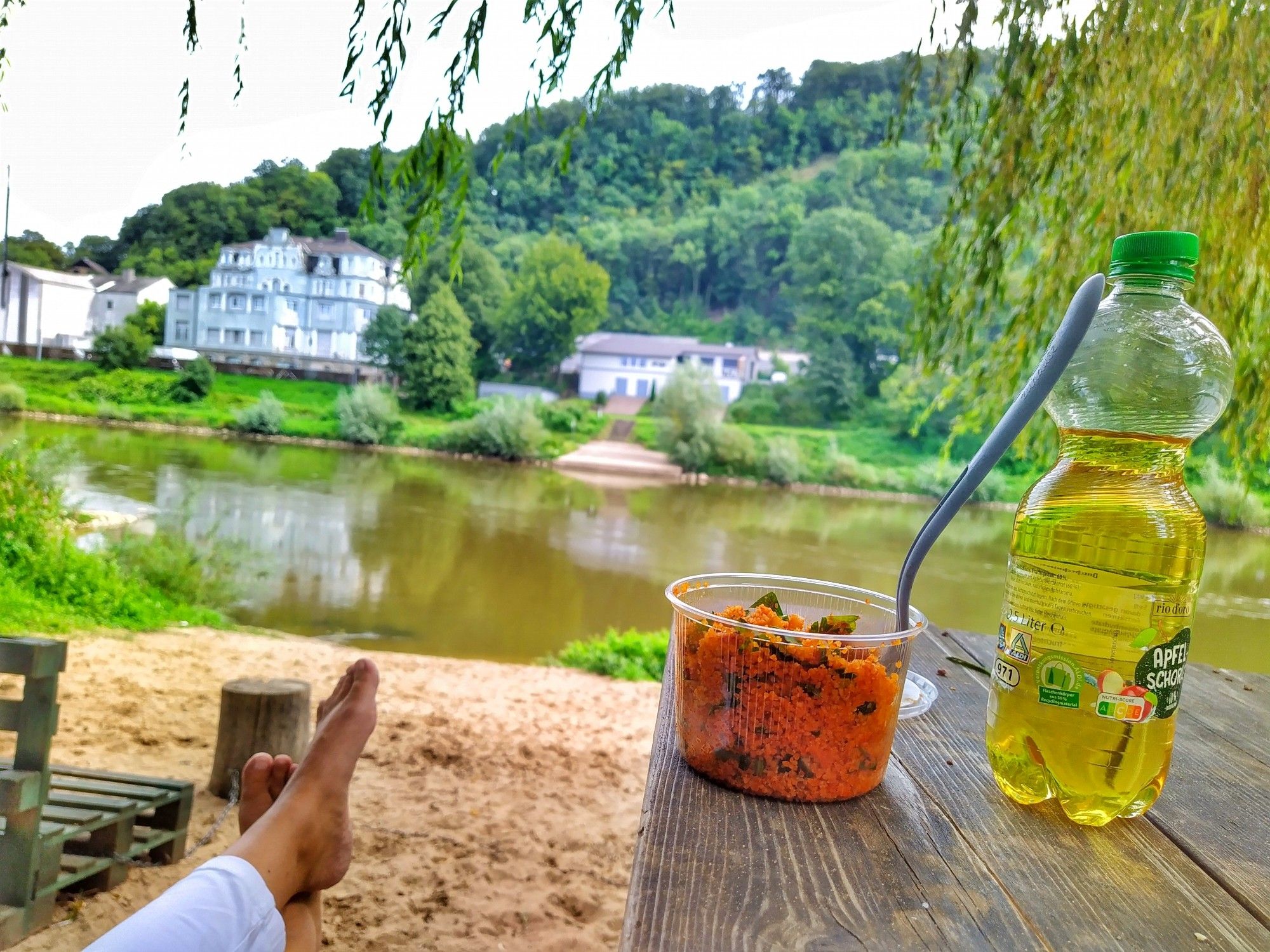 Auf einem hohen Holztisch steht ein Becher bulgursalat, eine Flasche apfelschorle, dahinter meine Beine mit nackten Füßen, samt einer Beachbar und die Weser im Hintergrund eine Villa aus dem 19 Jahrhundert