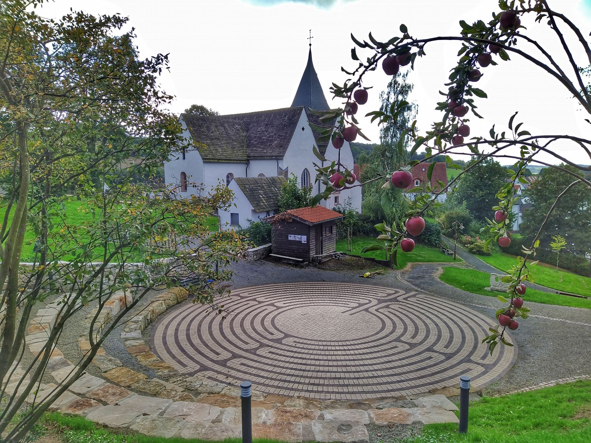 Ein aus Stein geschaffenes Boden Labyrinth mit Sitzplätzen aus Sandstein, dahinter weiße Kirche mit spitzen Turm