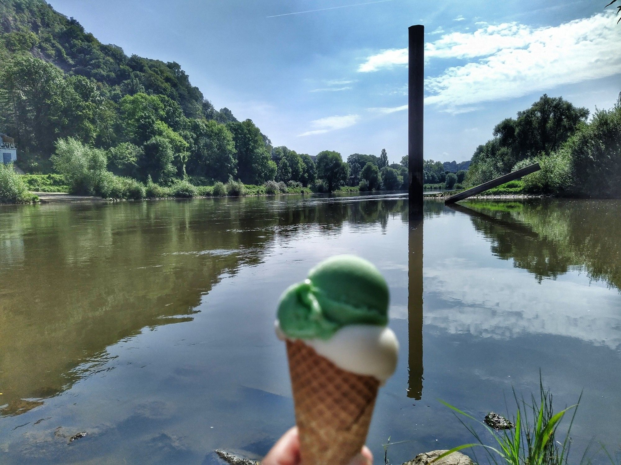 Eis mit zwei Kugeln in einer Waffel, dahinter die Breite glatte Visa, ein metallpfosten steckt mehrere Meter hoch im Wasser, bewaldetes Ufer