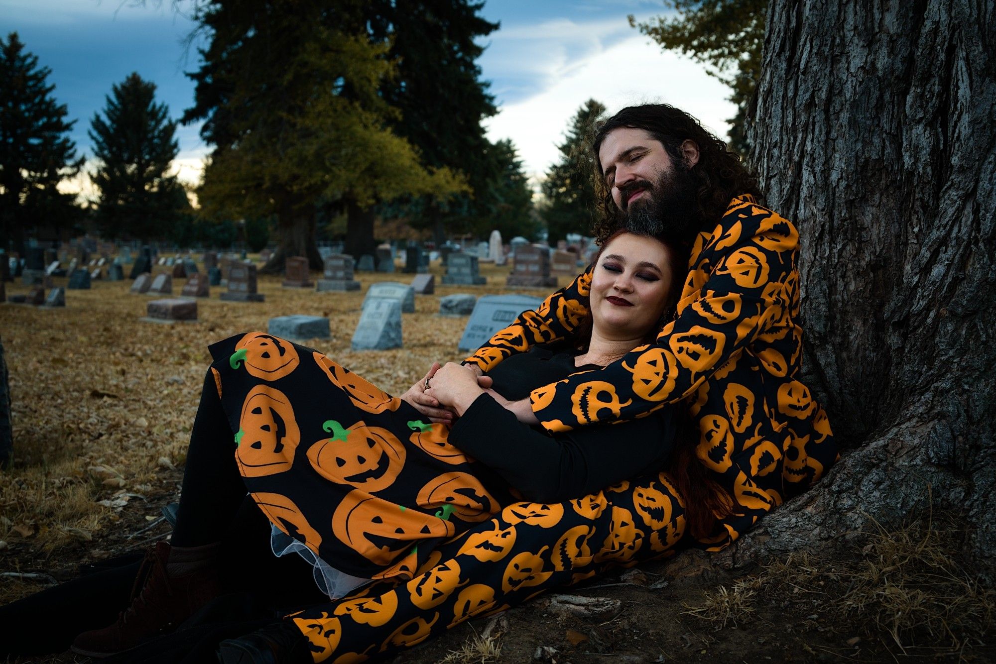 Jillian and Chris, wearing their pumpkin patterned formal wear, lay at the base of a tree in a graveyard and cuddle.